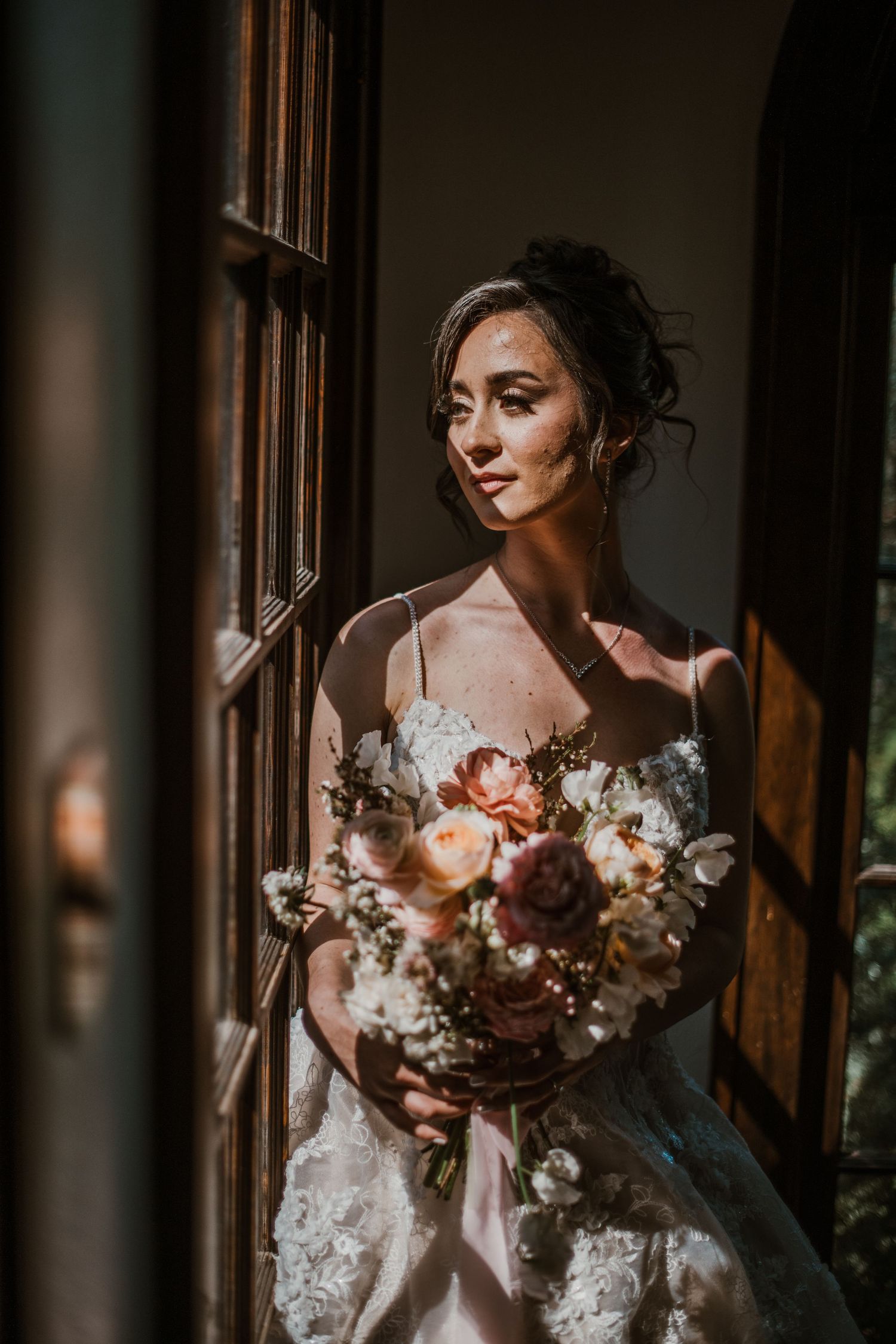 A bride in a white lace dress stands by a window holding a beautiful bouquet of pink and white flowers.