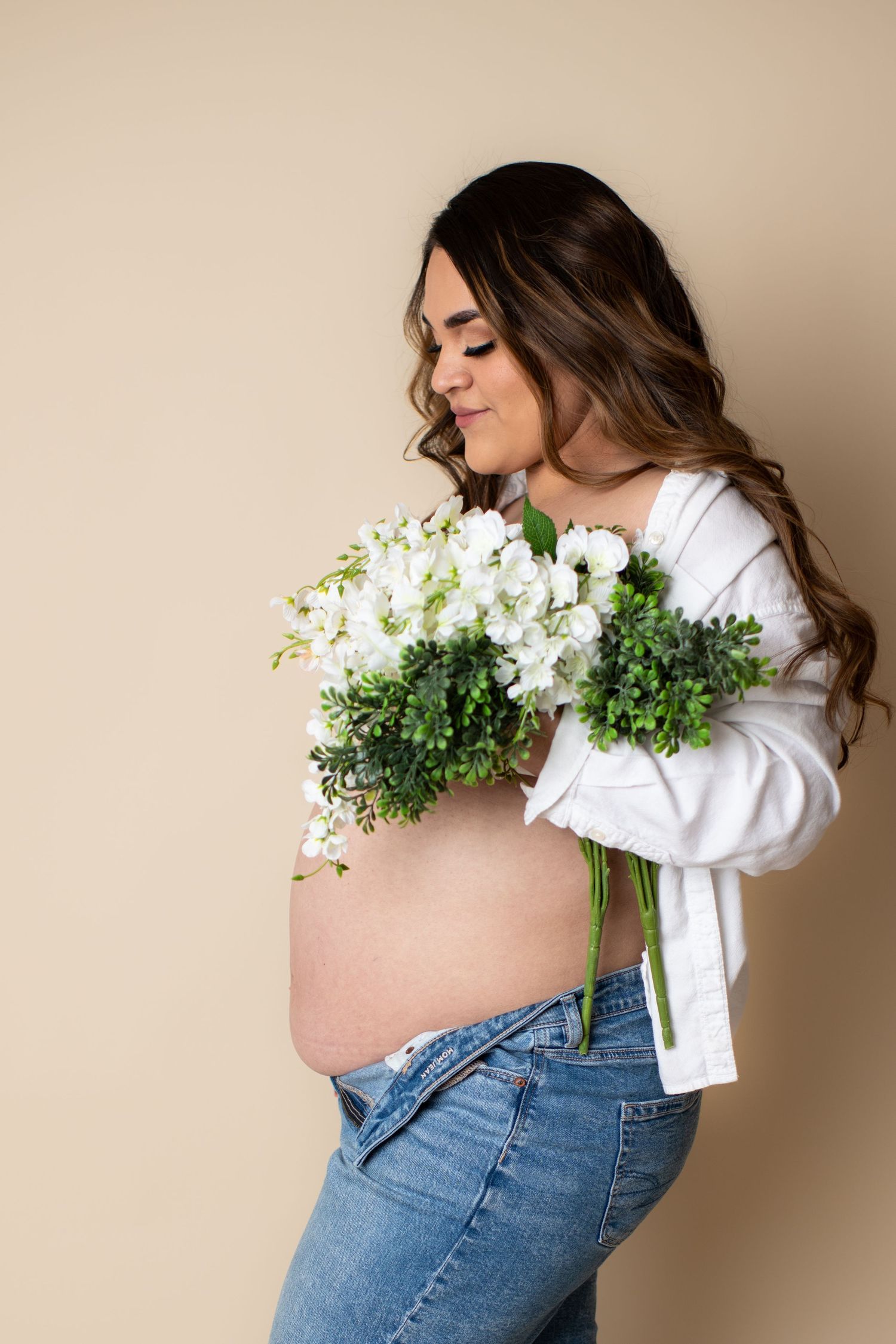 Expectant mother in white top and jeans poses with white floral bouquet against beige background during maternity shoot.