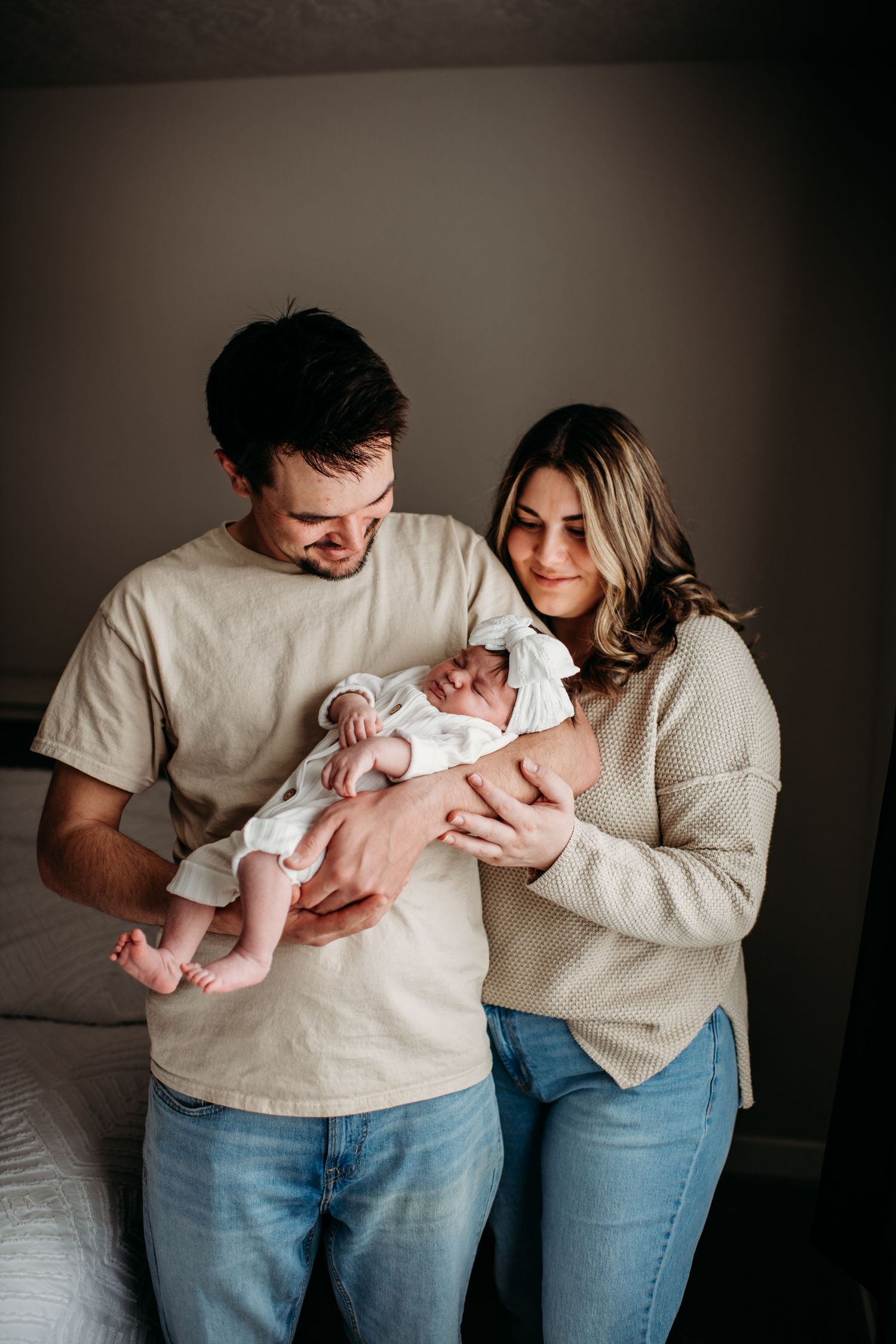 A couple in casual attire tenderly holds their newborn baby in a dimly lit room.