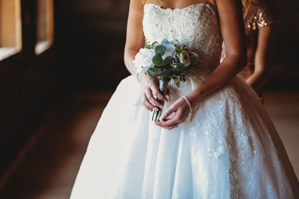 Close up of a bride in a white lace ballgown holding a small bouquet with blue flowers and greenery.