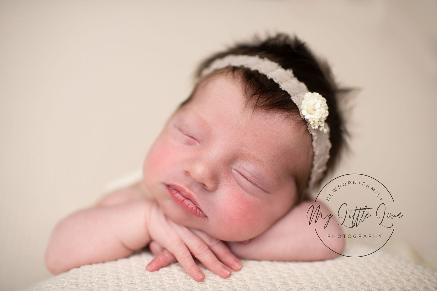 A sleeping newborn baby wearing a delicate lace headband rests on a soft cream-colored fabric.