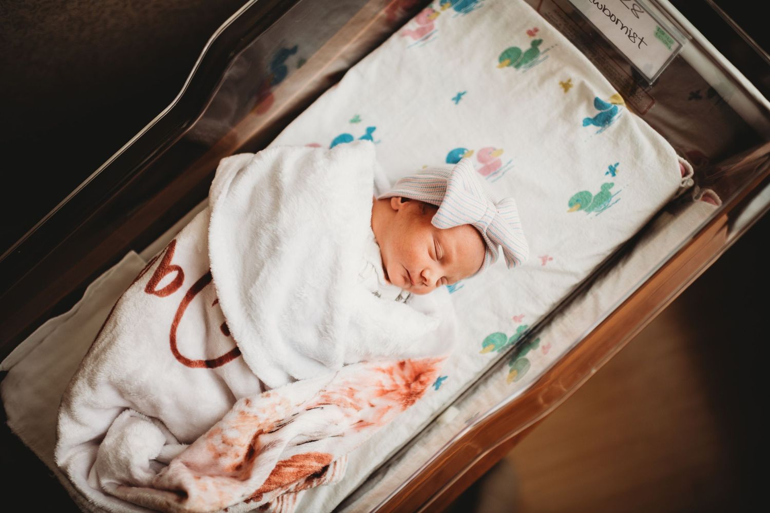 Newborn baby swaddled in white blanket with teal and green designs lying in hospital bassinet.