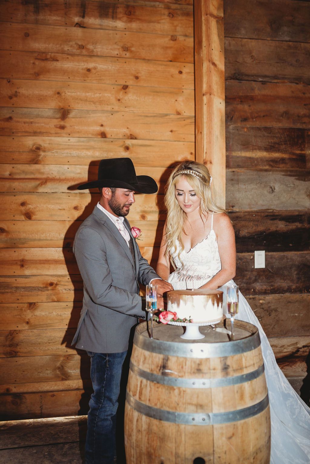 Couple cuts wedding cake together on rustic wooden wine barrel in barn venue with wooden plank walls.