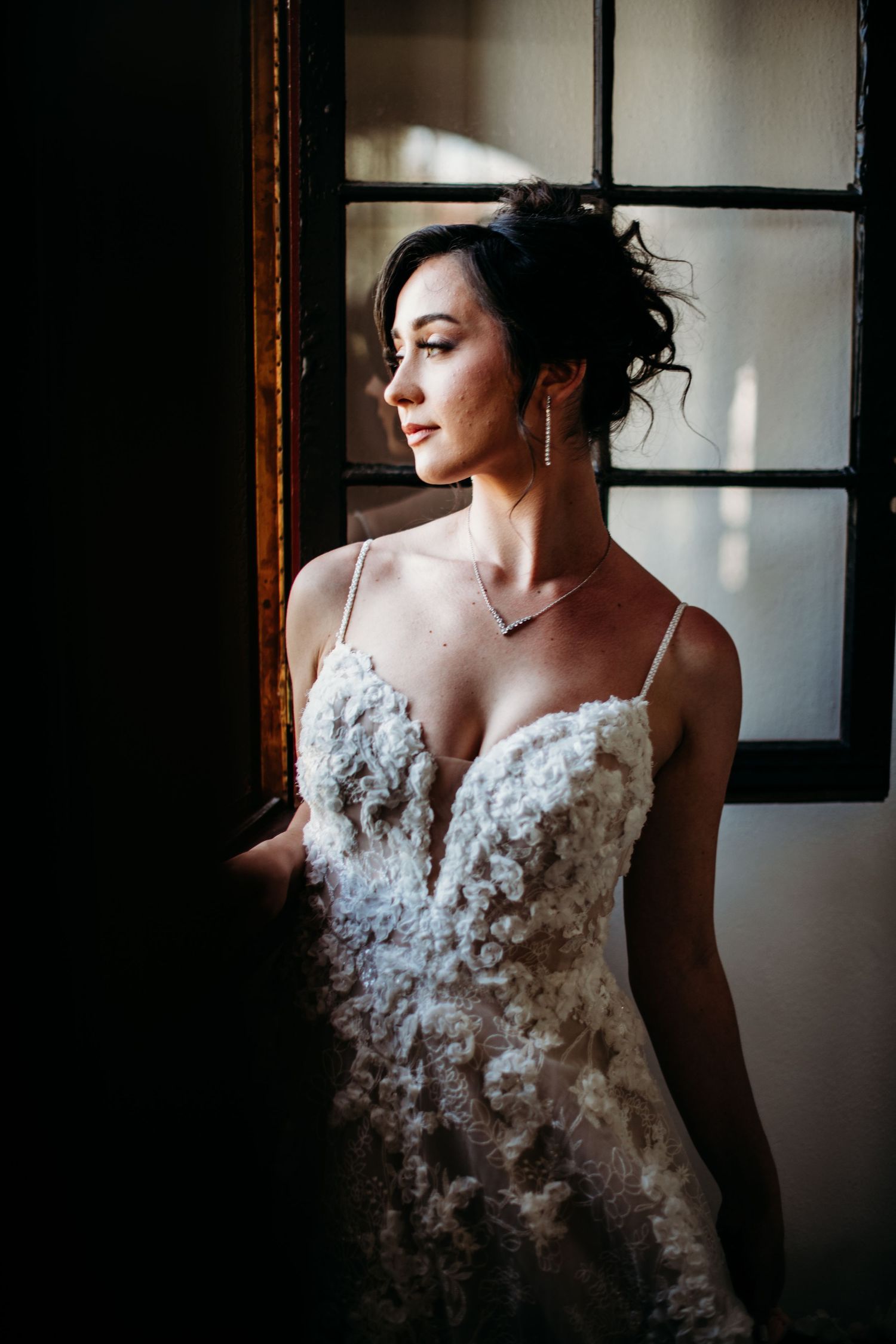 A bride in an elegant white lace wedding dress with spaghetti straps poses near a window in moody natural light.