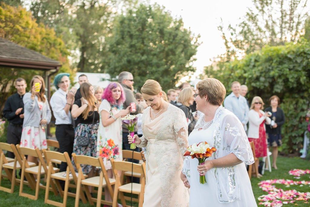 Guests gather at an outdoor wedding ceremony with wooden chairs and floral arrangements on a sunny day.