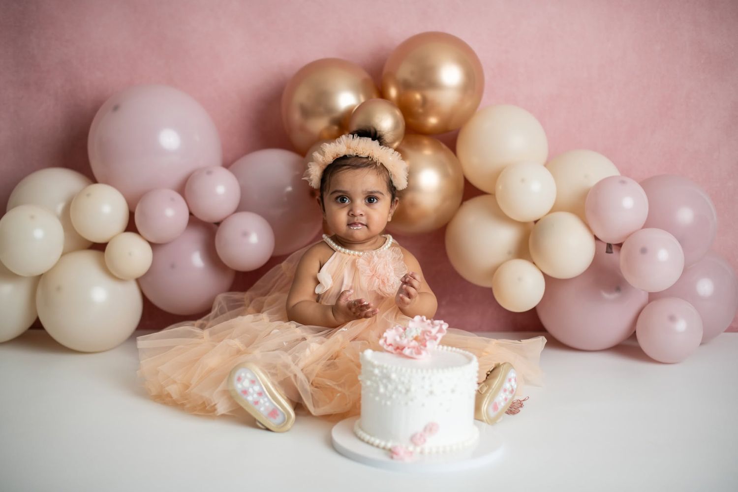 Baby girl in peach dress sits before a white cake surrounded by pink and gold balloons for her first birthday smash cake photo.