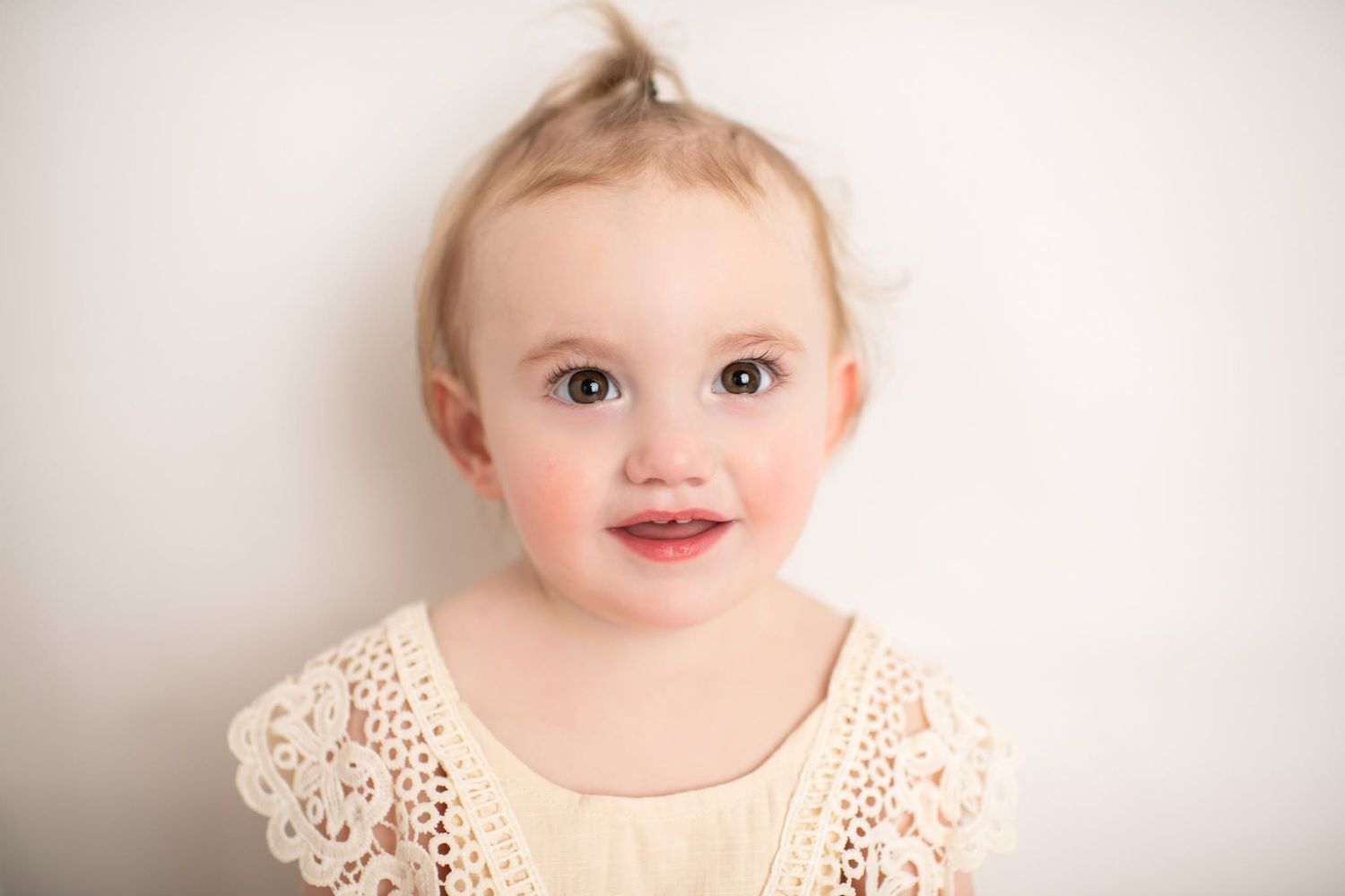 Adorable toddler girl with rosy cheeks and a tiny ponytail smiling sweetly in a cream lace dress.