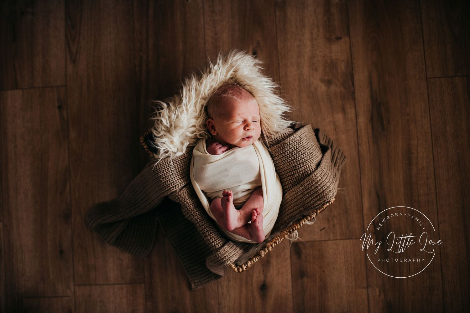 A newborn baby sleeps in a cozy brown basket on a wooden floor with fur lining.