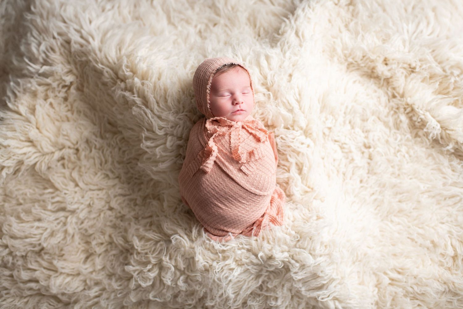 Sleeping newborn wrapped in soft pink fabric lies peacefully on fluffy white blanket during professional photo session.