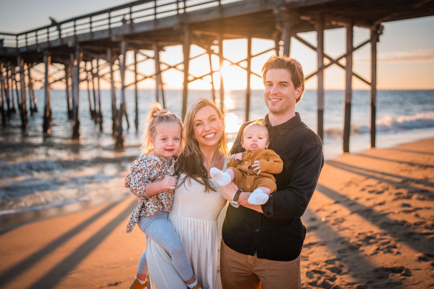 A group poses together on a beach at sunset with a wooden pier in the background.