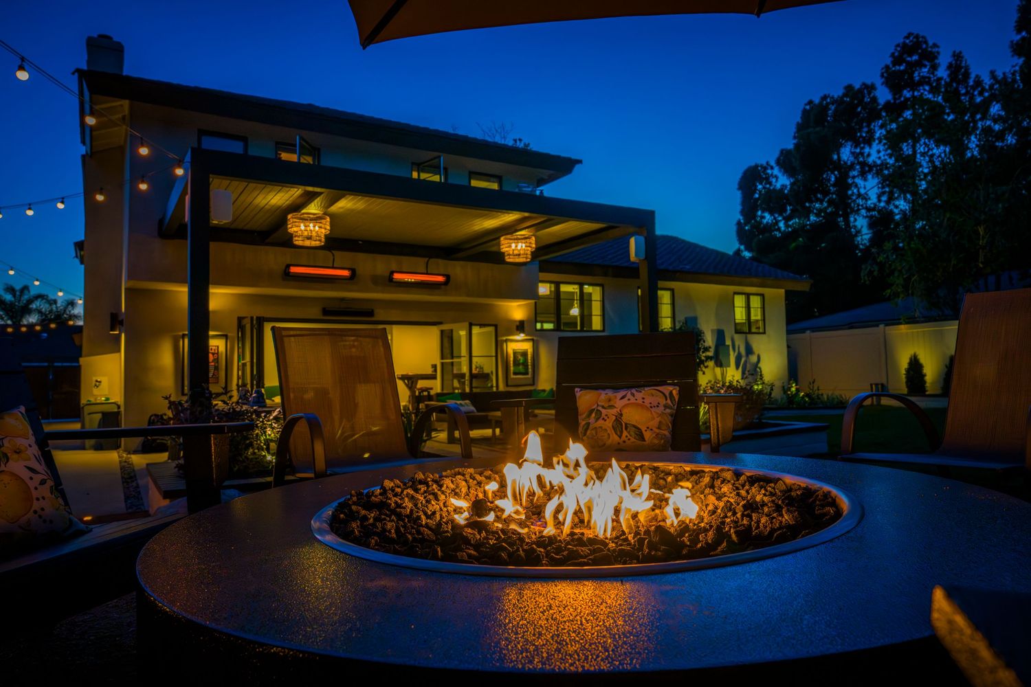 Modern house exterior with large circular fire pit glowing at night under string lights.