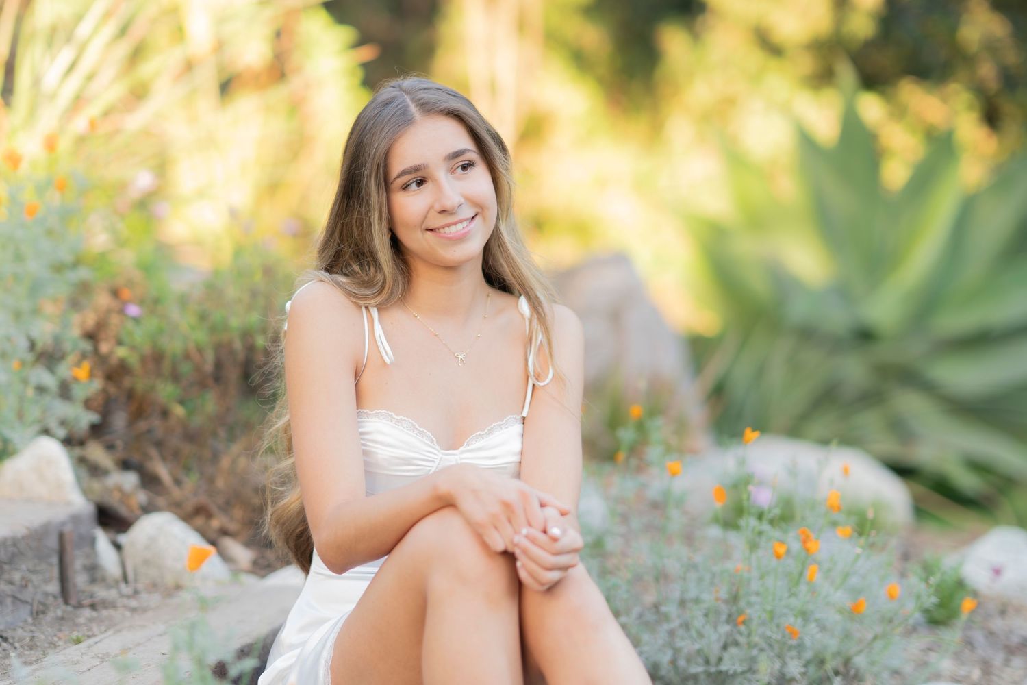 Young woman in white dress sitting outdoors among rocks and wildflowers, smiling in a sunny garden setting.