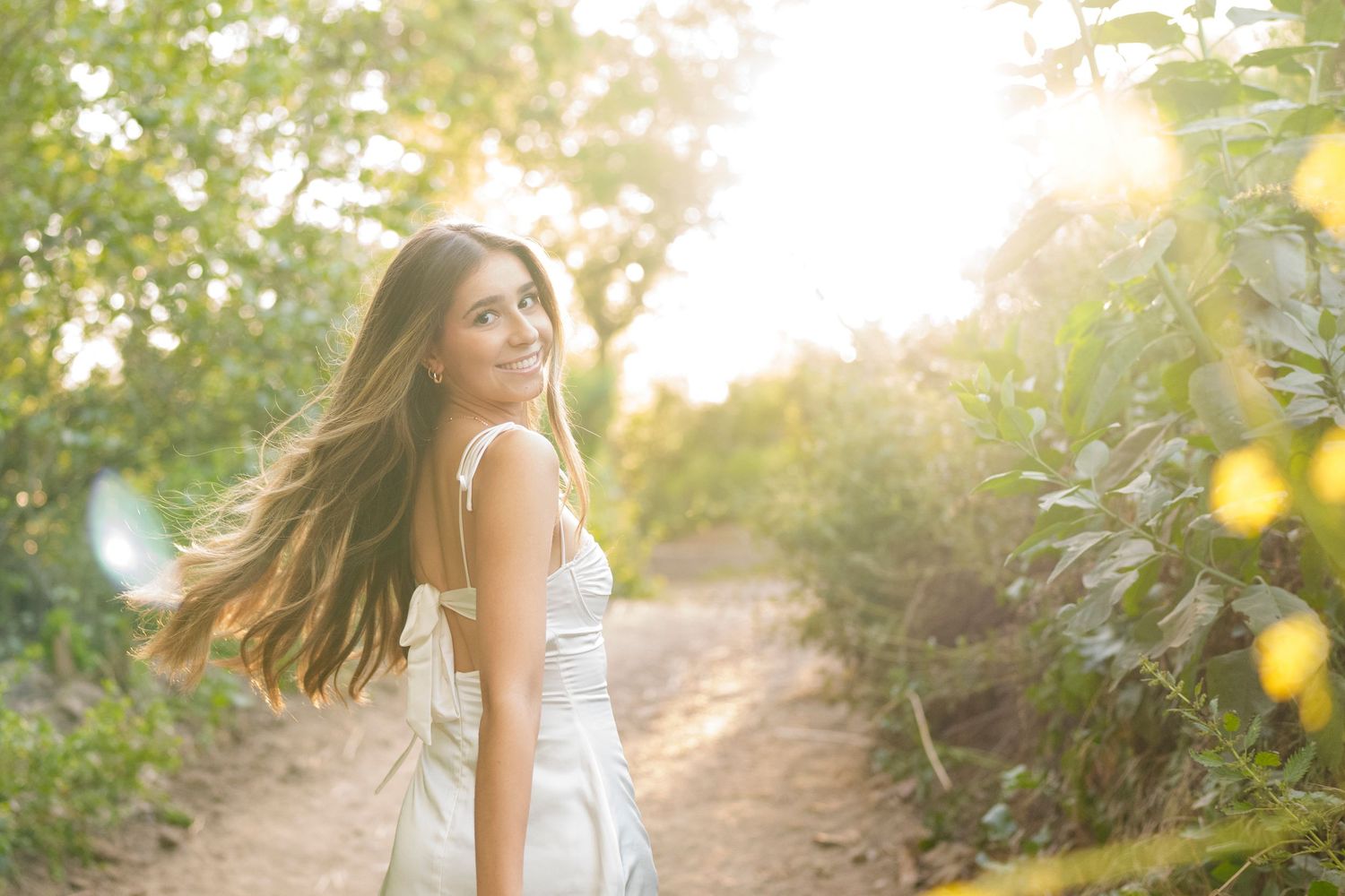 Young woman in white dress smiling over shoulder on sunlit forest path with long flowing hair.