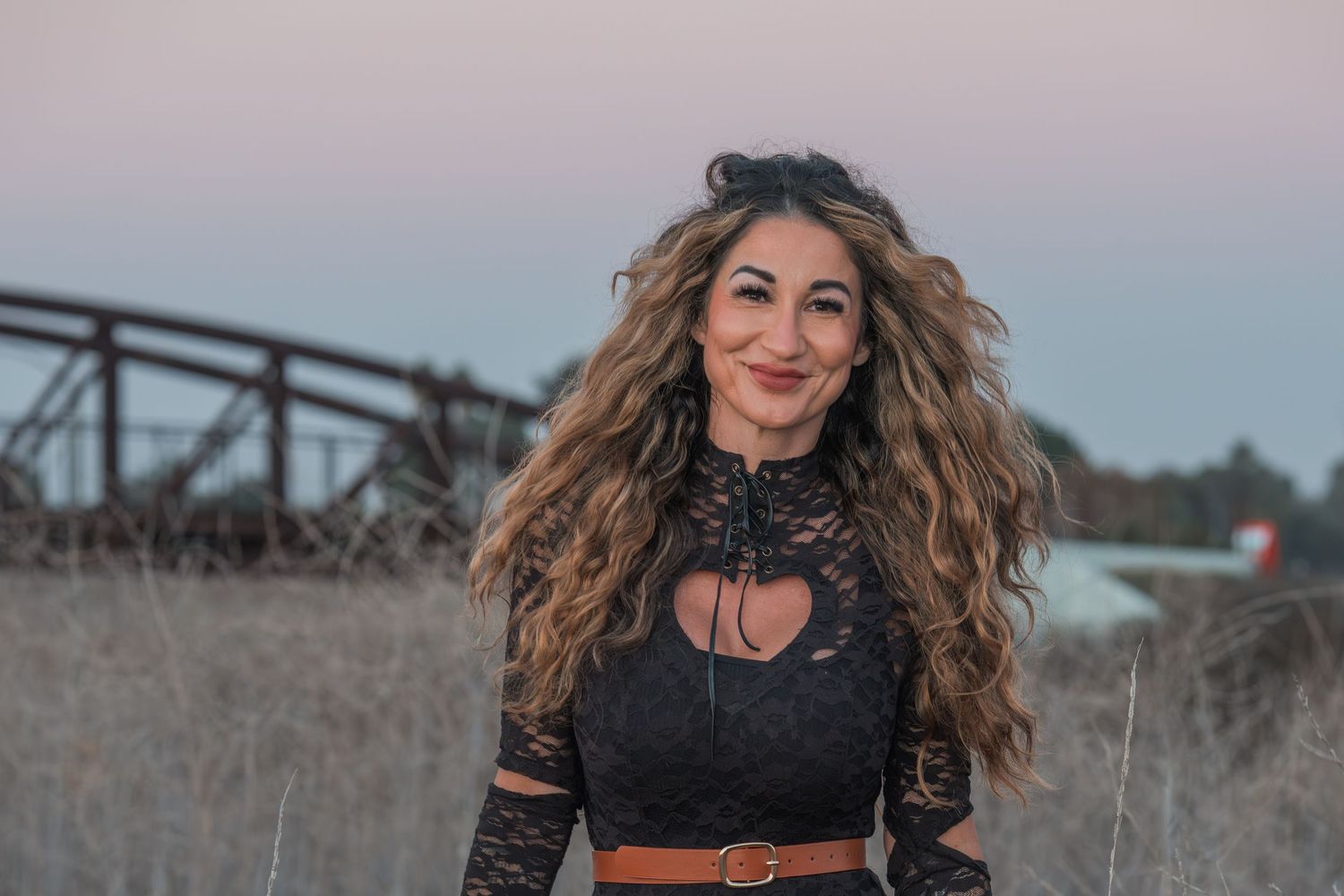 Long wavy hair blows in the wind against a sunset sky with an industrial bridge structure in the background.