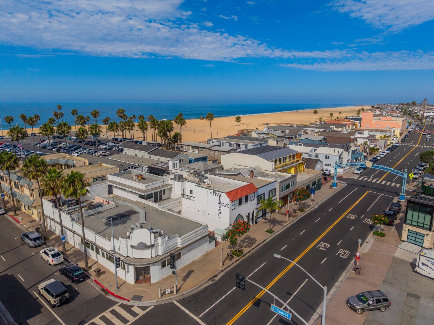 Aerial view of a coastal California town with palm trees, shops, and a sandy beach under a bright blue sky.