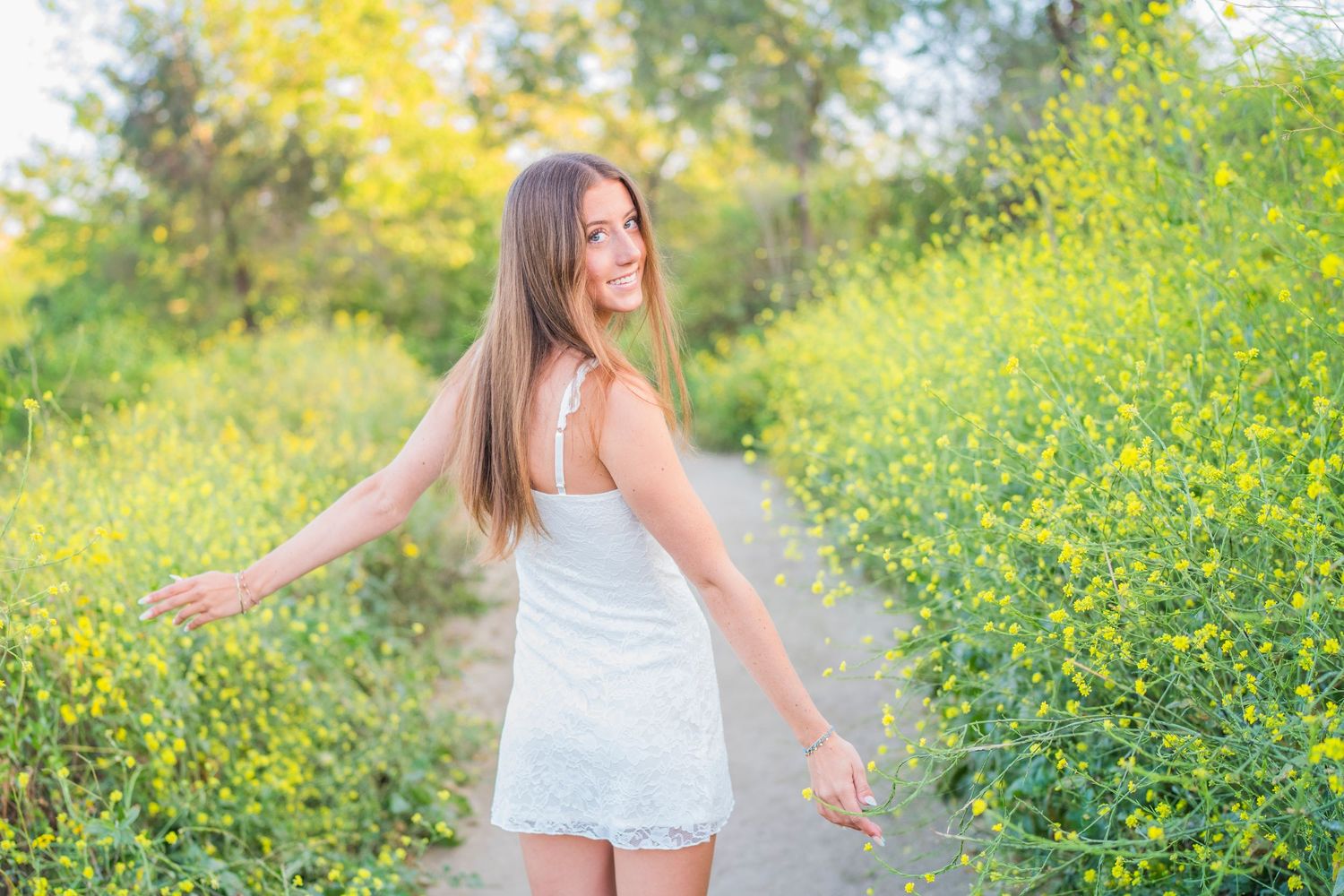 Young woman in white dress smiling on a path surrounded by yellow wildflowers on a sunny day.
