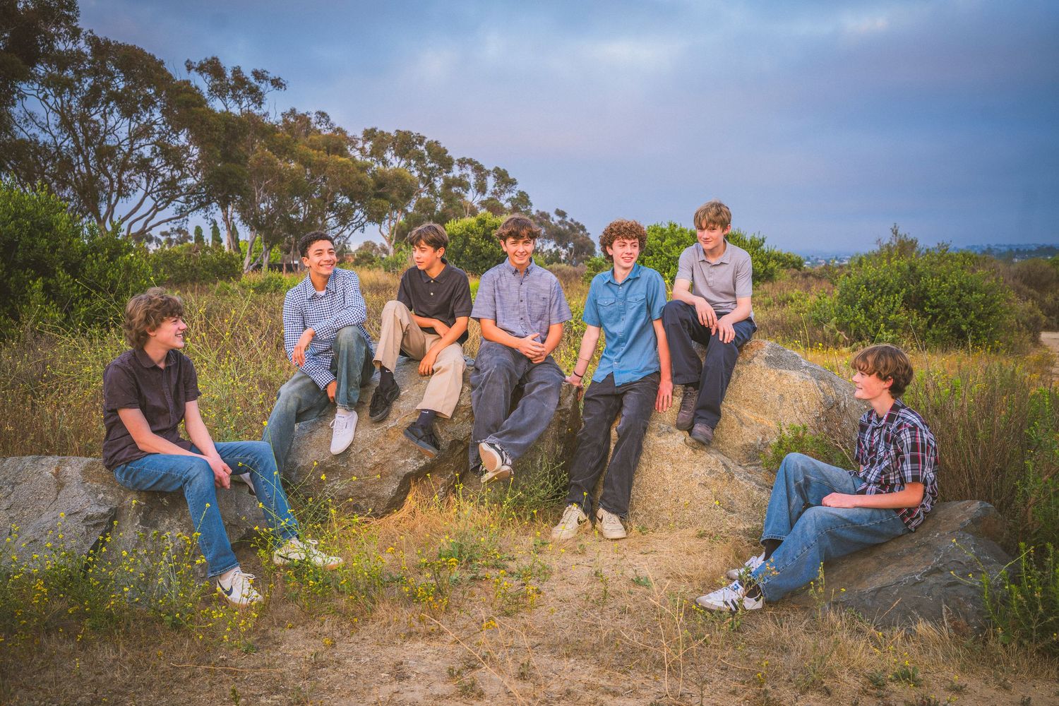 Seven teenage boys sitting on rocks in a natural outdoor setting with green hills and trees in the background.