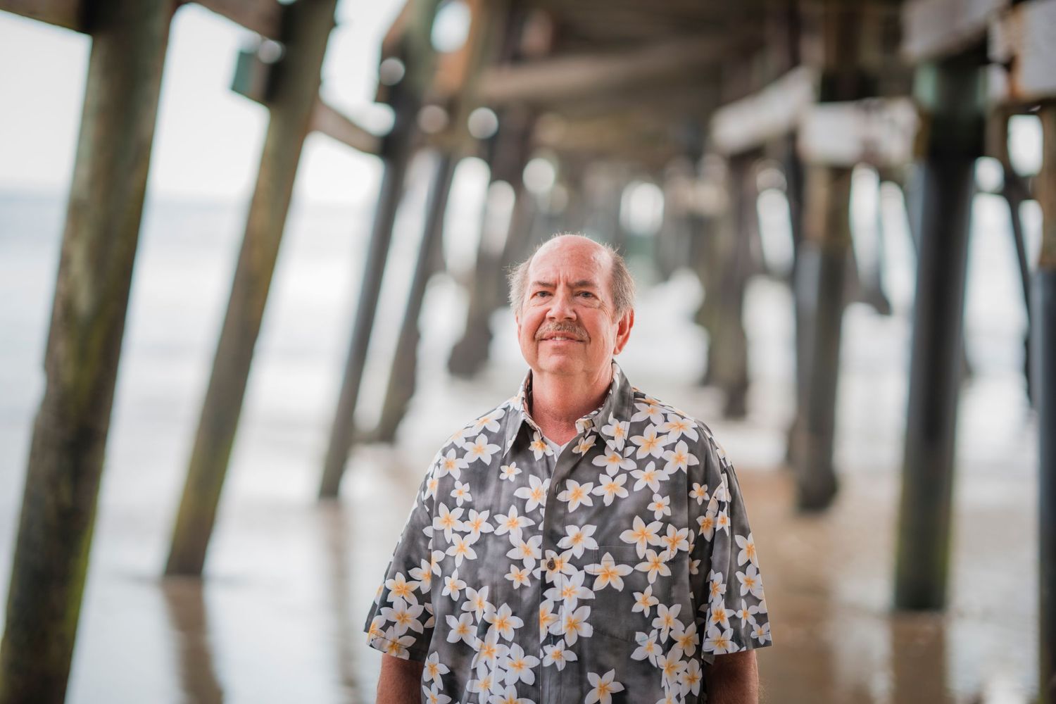 A couple in floral shirts shares a joyful laugh together while posing near wooden pilings on a beach.