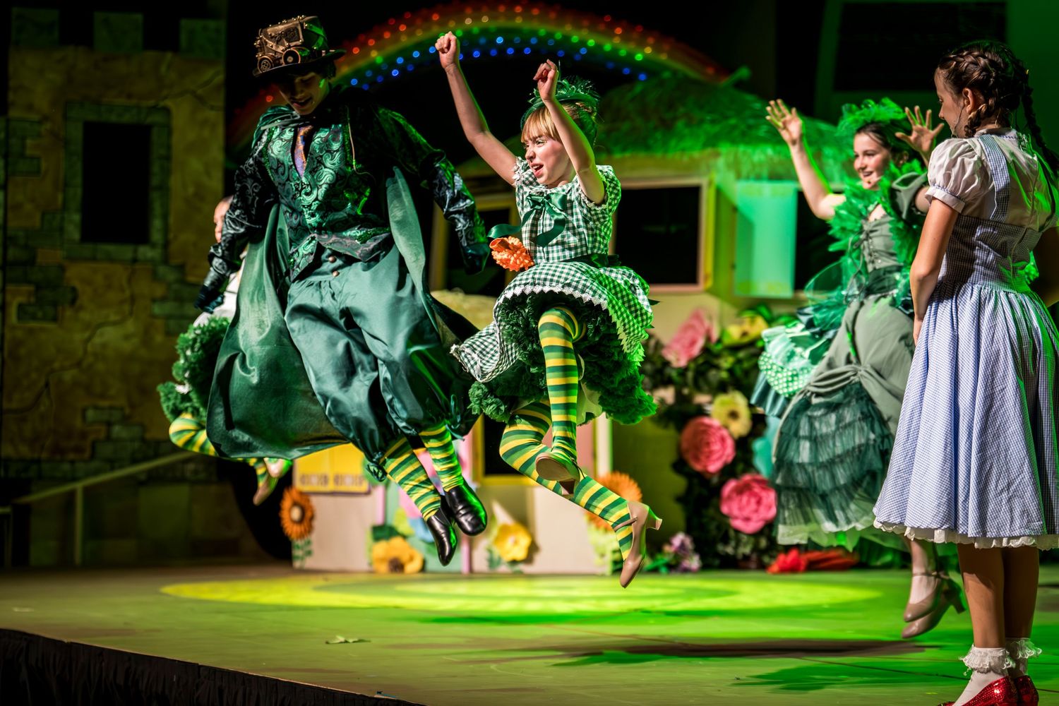 Stage performers dancing under green lighting with a rainbow backdrop in a theatrical show.
