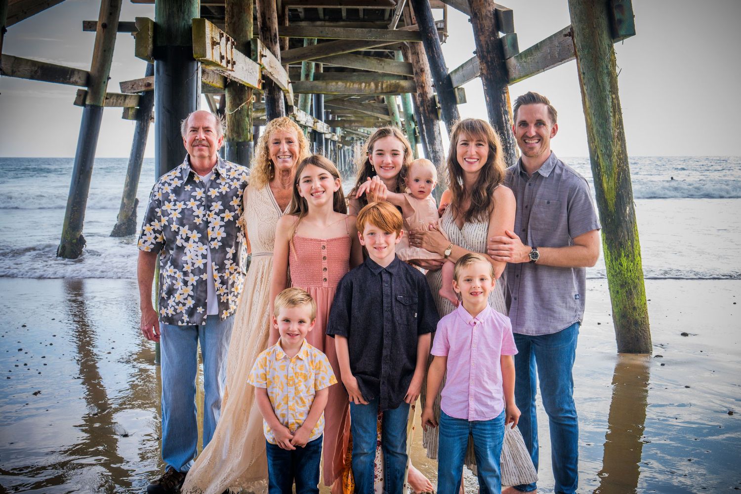 A family poses together on a beach boardwalk with wooden pilings and ocean waves visible in the background.