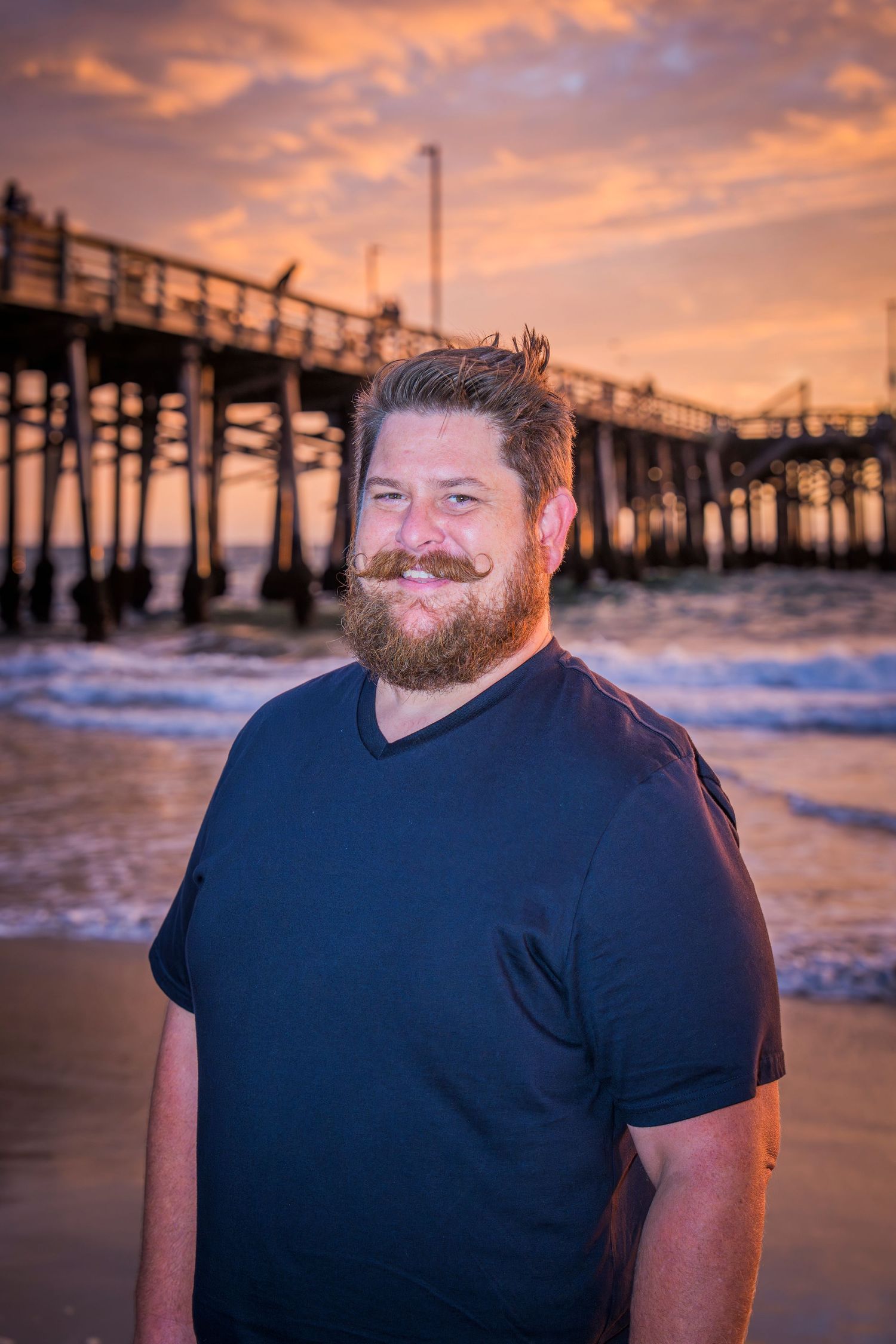 A bearded man in a navy t-shirt stands on a beach at sunset with a wooden pier in the background.