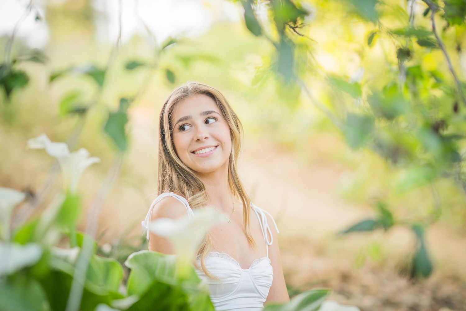 Smiling young blonde woman in white dress surrounded by lush green leaves and white flowers in a sunlit garden.