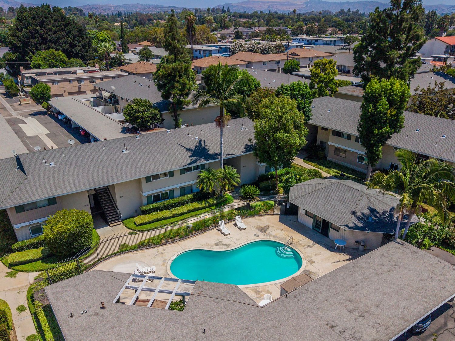 Aerial view of a residential apartment complex with a kidney-shaped pool surrounded by lounge chairs and lush trees.