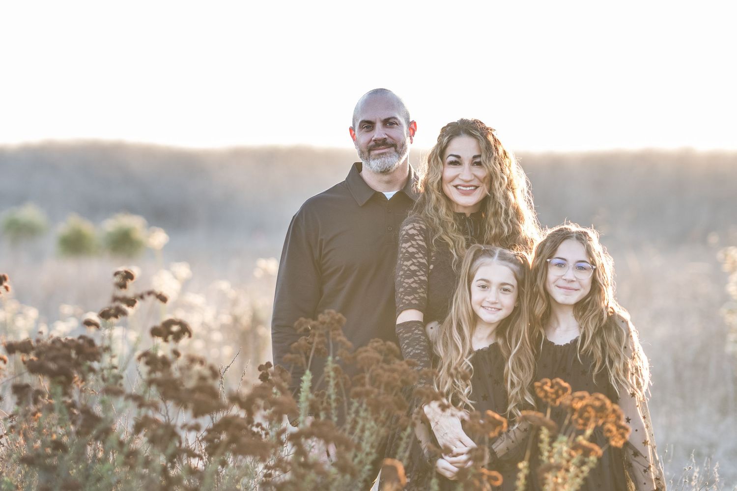 Family members laugh together while standing in a sunlit field during golden hour at sunset.