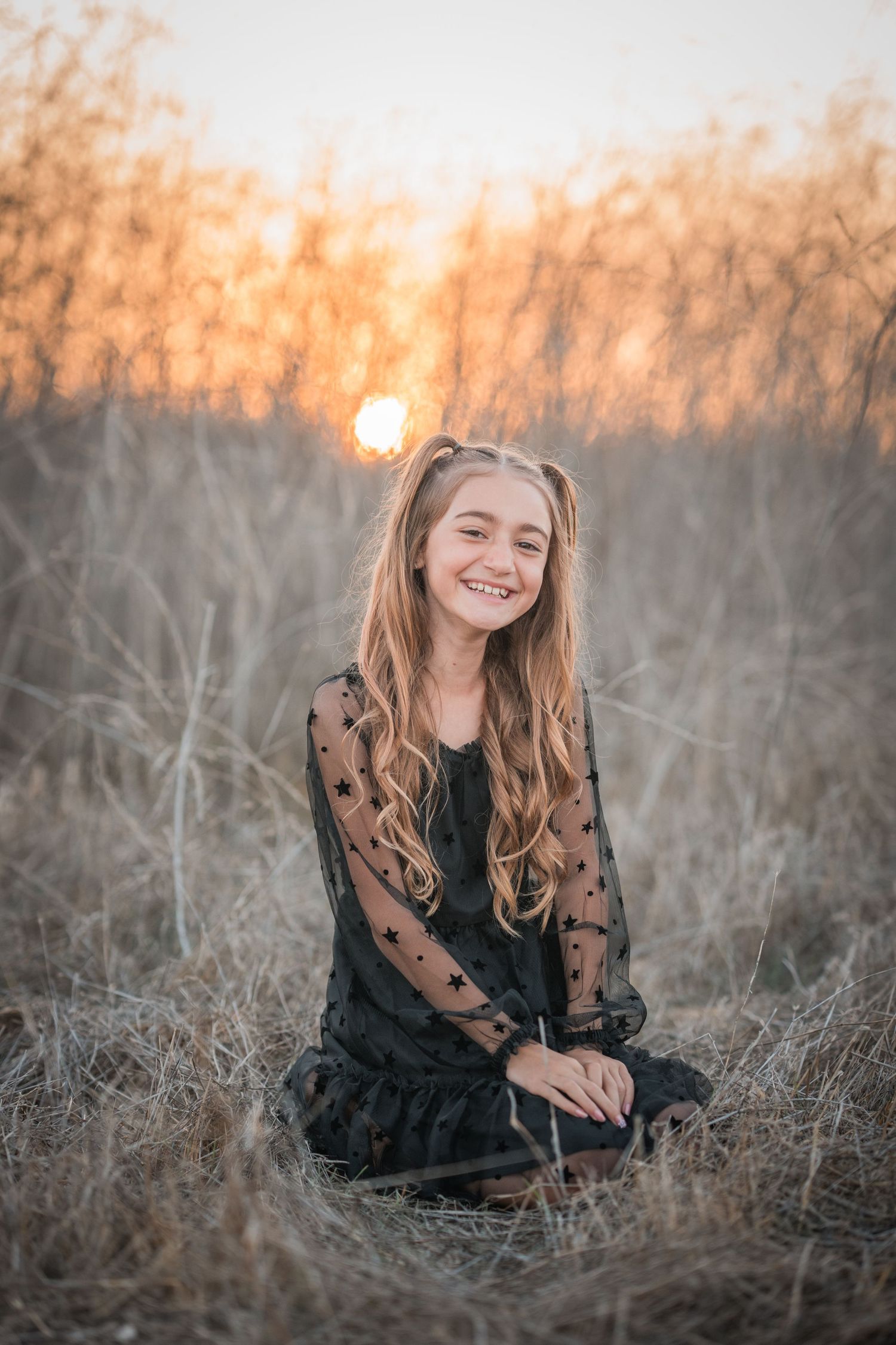 A person in a black dress sits in a field during golden hour sunset with tall dried grass and warm lighting.