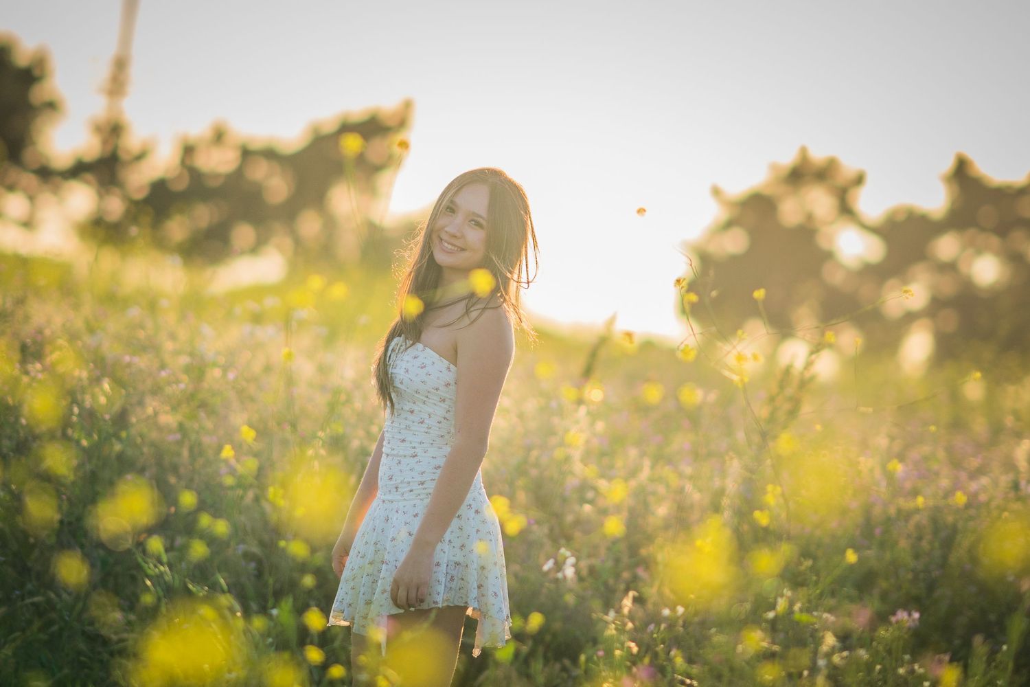 Young woman in white dress smiling in a sunlit wildflower meadow with golden bokeh and lush green trees.
