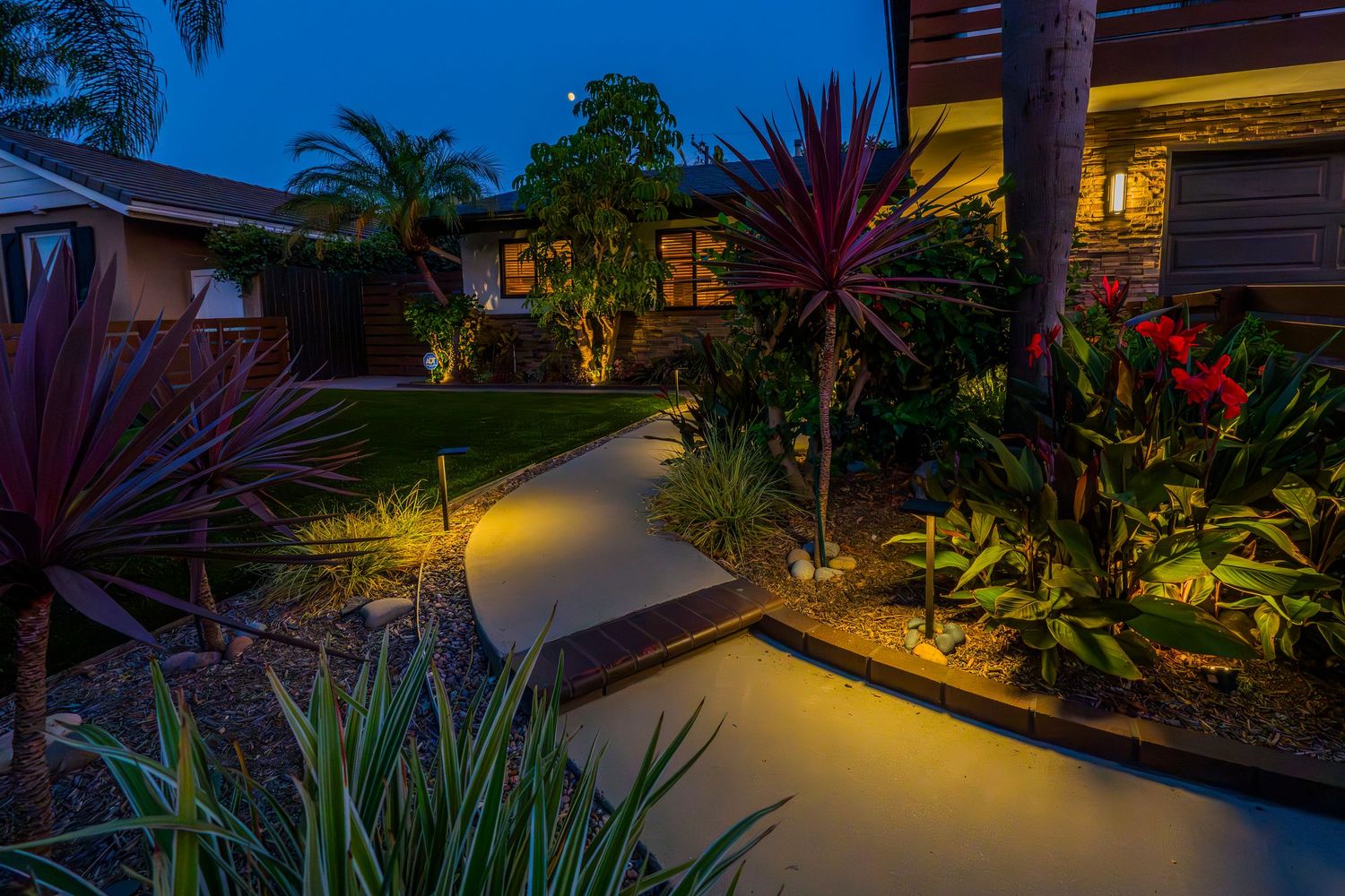 Evening garden landscape with illuminated walkway and tropical plants under blue twilight sky.