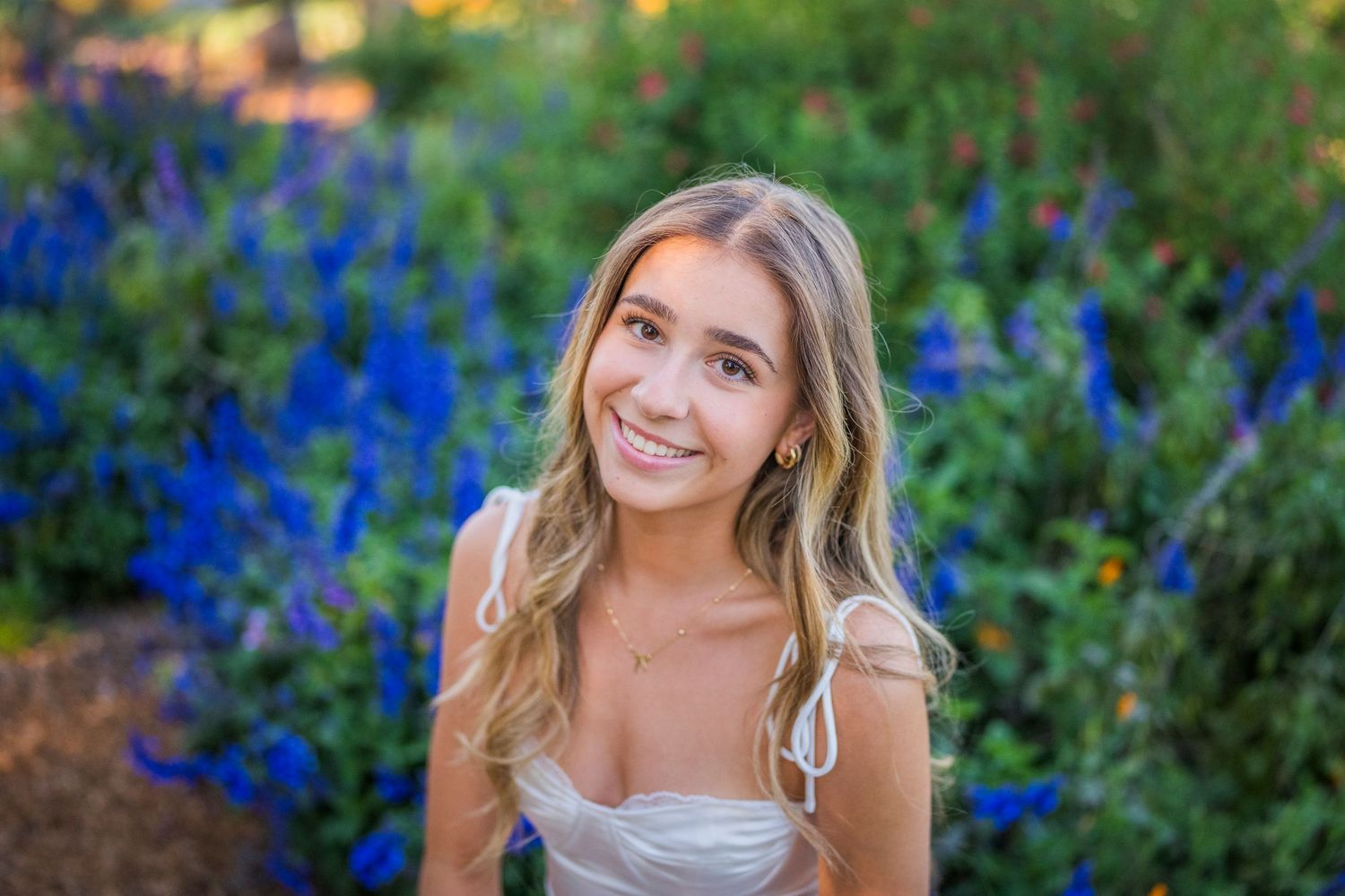 Smiling teenage girl with long blonde hair posing outdoors in front of vibrant blue and purple flowers.