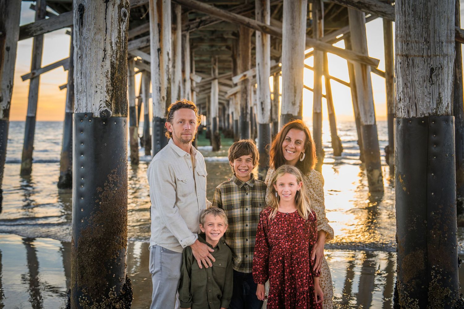 Family photo session under wooden pier at sunset with ocean waves in background.