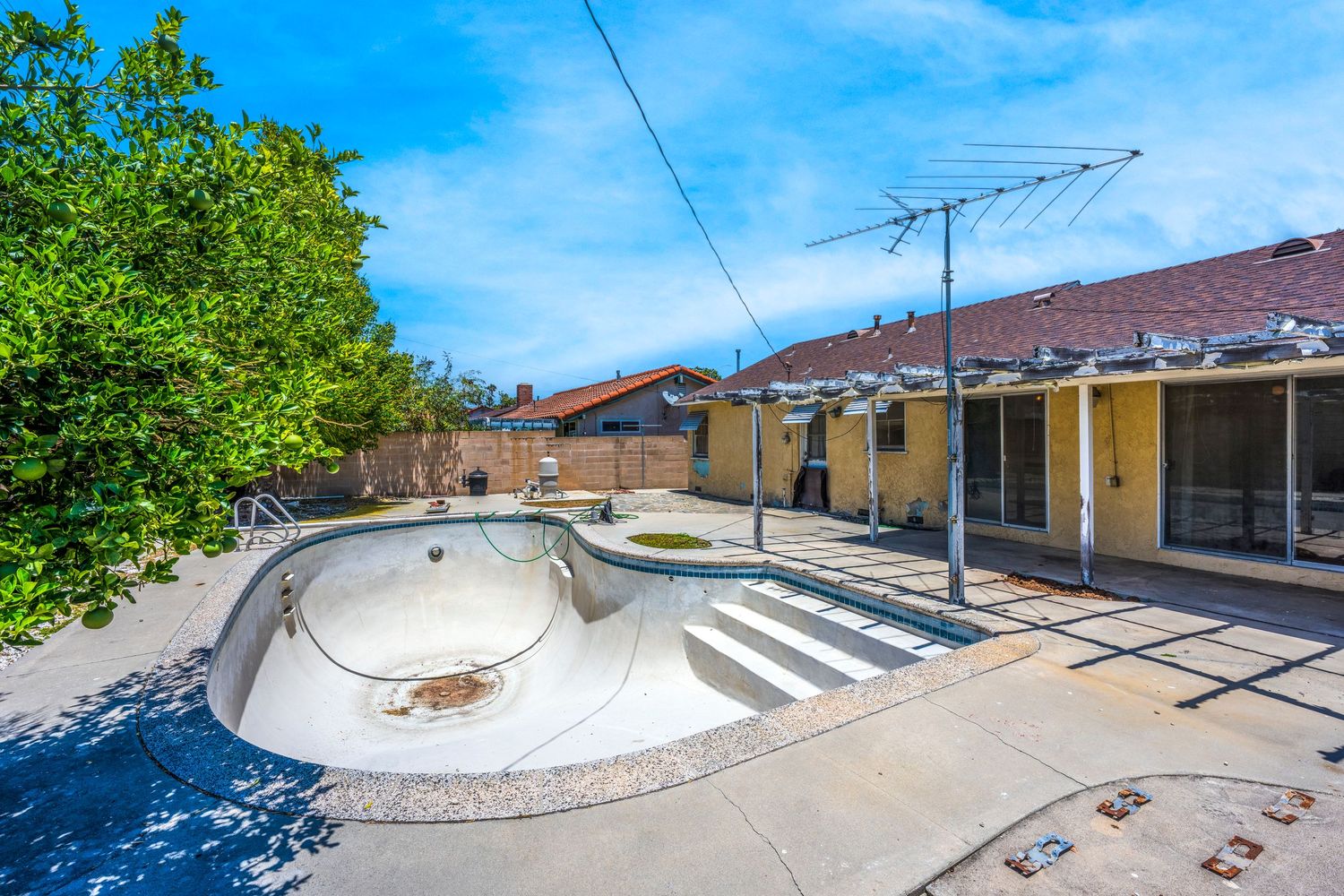 Empty drained swimming pool in a sunny backyard patio with surrounding concrete deck and residential home exterior.