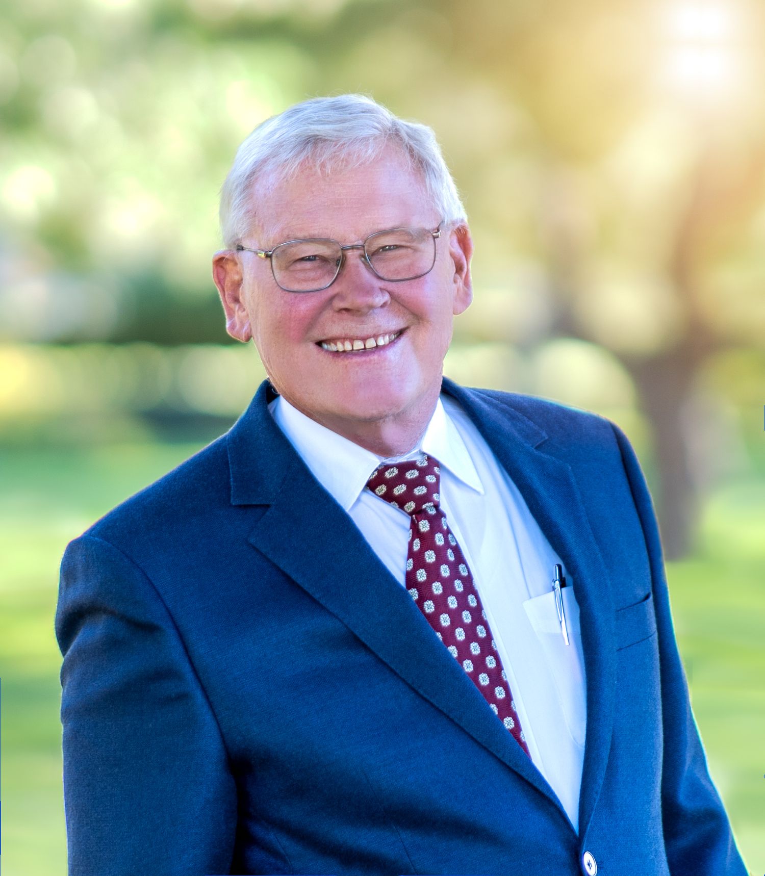 Professional headshot of person in navy suit and red tie with outdoor background.