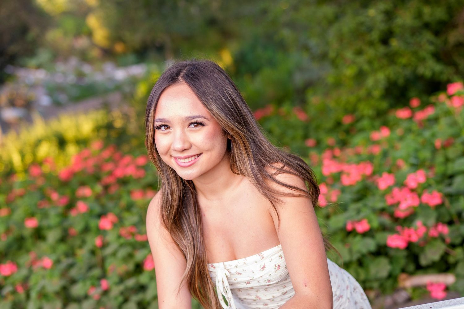Young woman smiling in a garden with vibrant pink flowers and green foliage in the background.