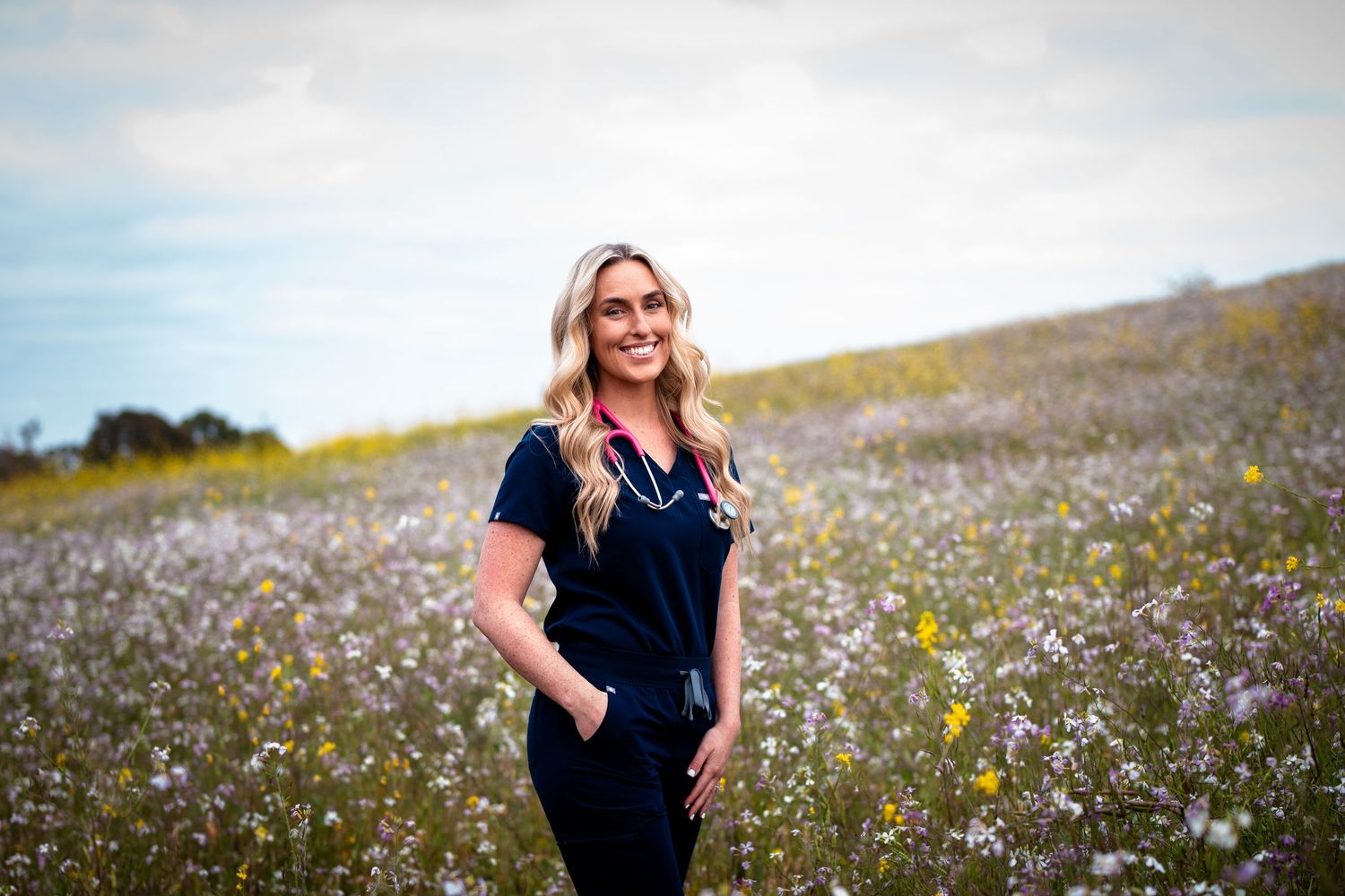 Smiling blonde woman in navy scrubs with stethoscope standing in colorful wildflower meadow under cloudy sky.