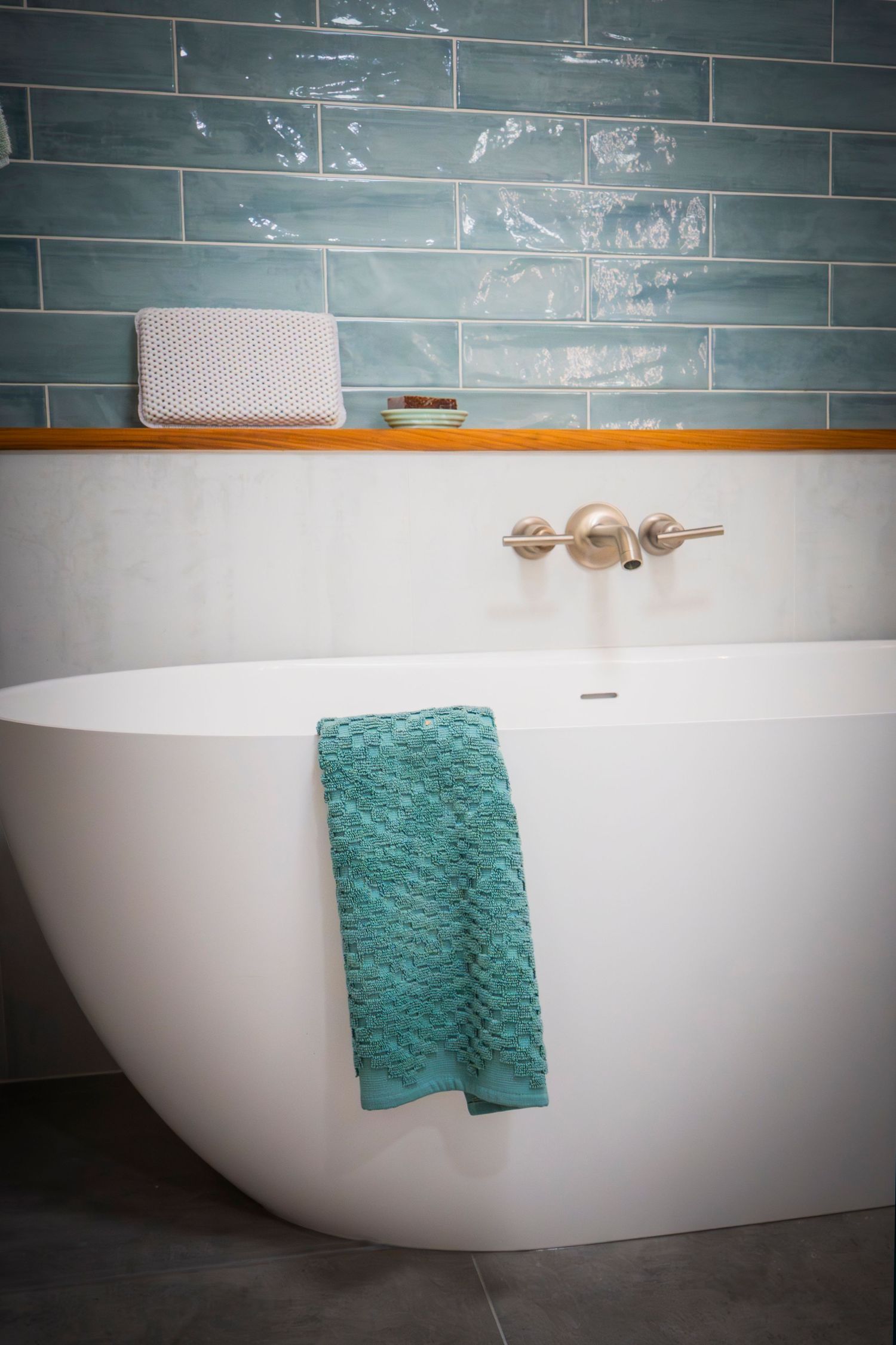 White freestanding bathtub with teal towel, gold fixtures, wooden shelf, and teal subway tile wall in modern bathroom.