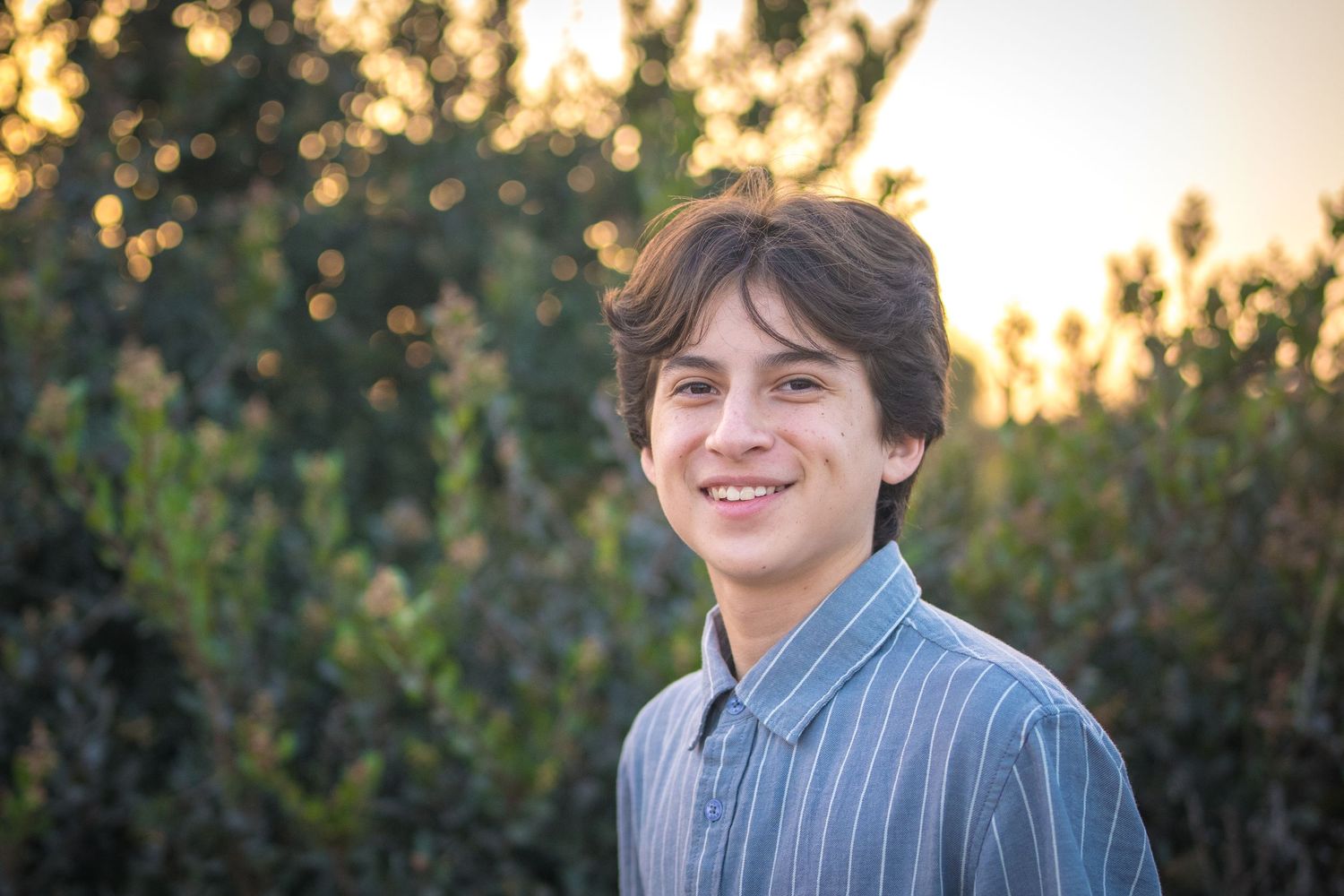 A young person in a striped shirt smiles while standing outdoors during golden hour sunset.
