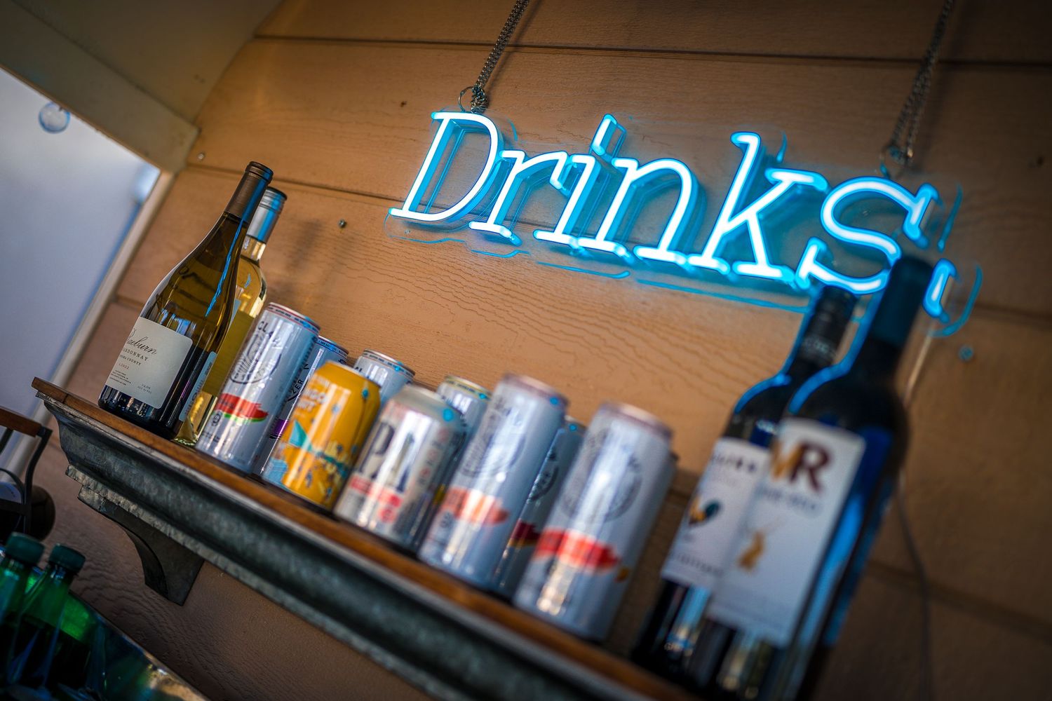 Blue neon 'Drinks' sign above assorted cans and bottles on wooden shelf.