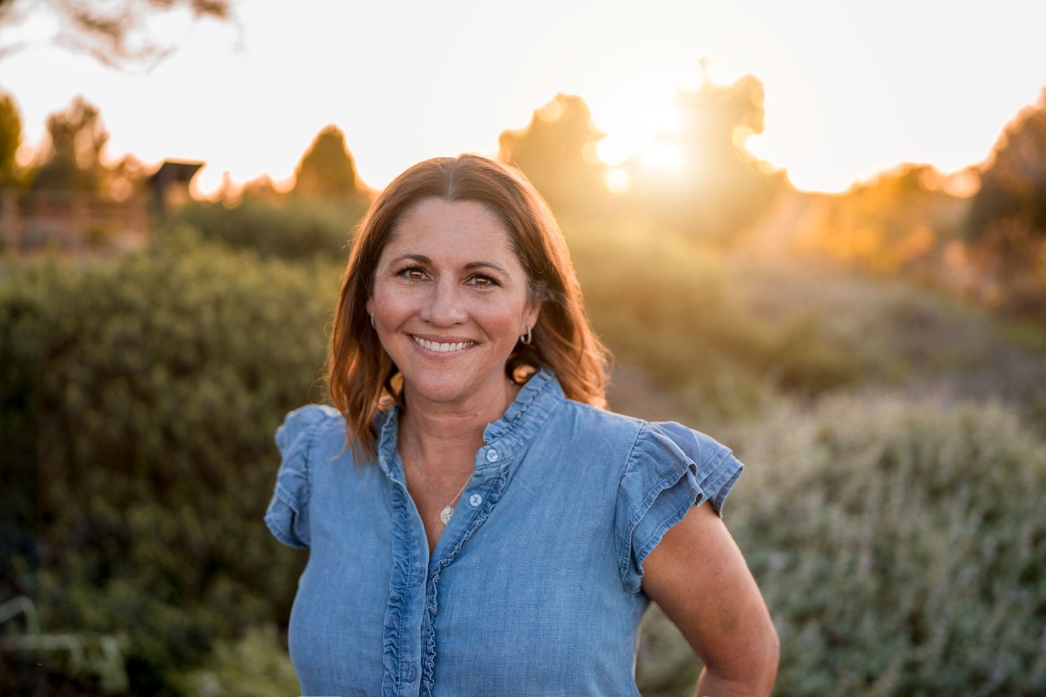 Woman in denim shirt standing outdoors during golden hour sunset.