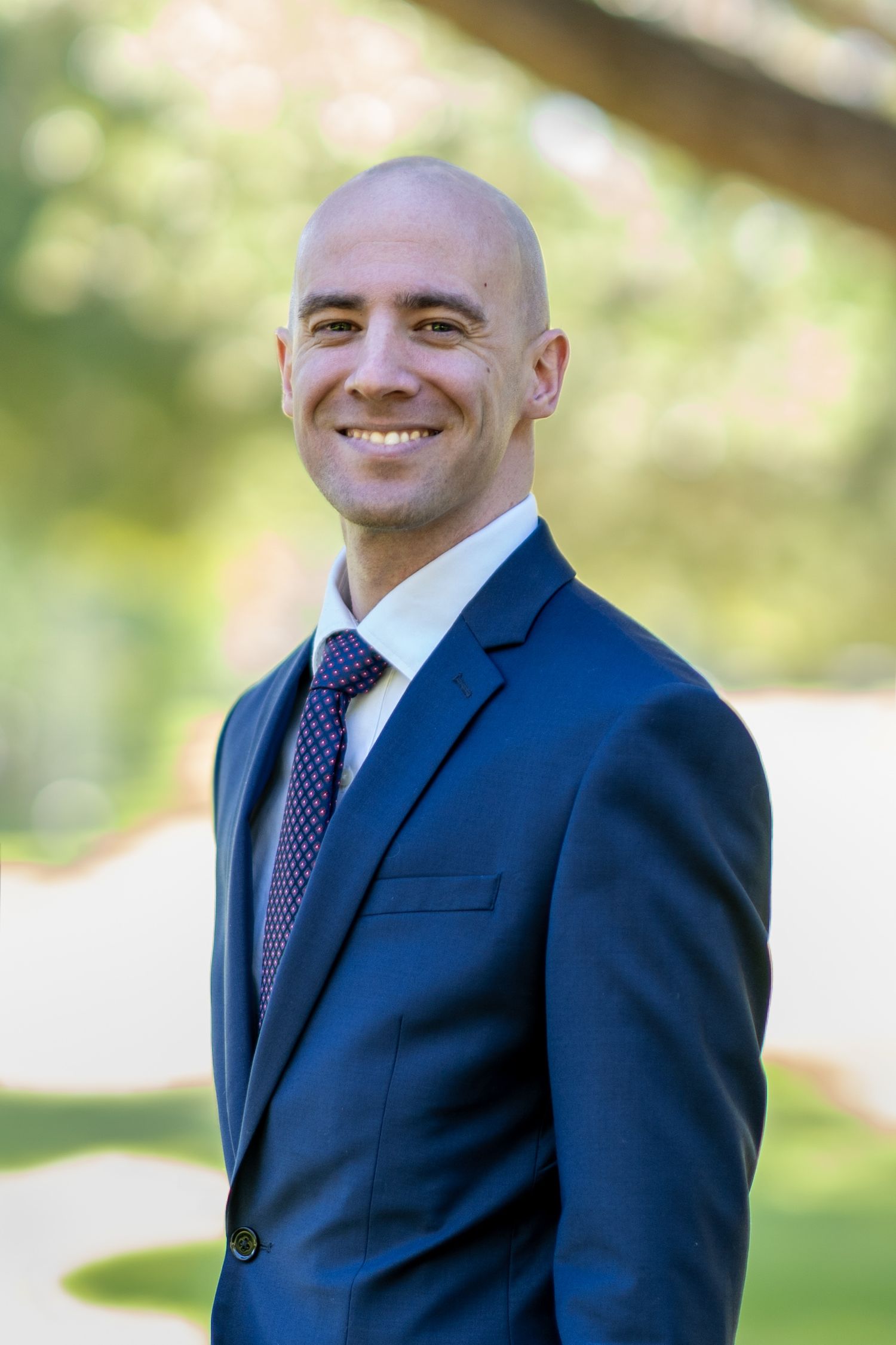 Corporate portrait in dark suit against blurred natural background.
