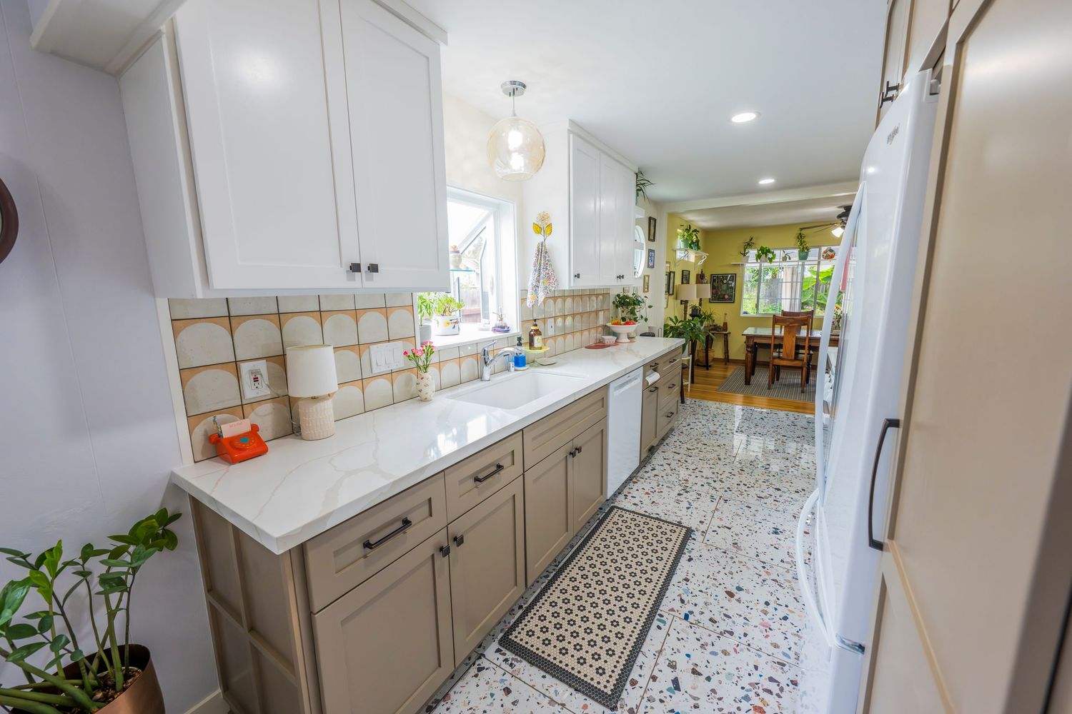 Bright galley kitchen with white cabinets, quartz countertops, terrazzo floors, and tile backsplash leading to dining area.
