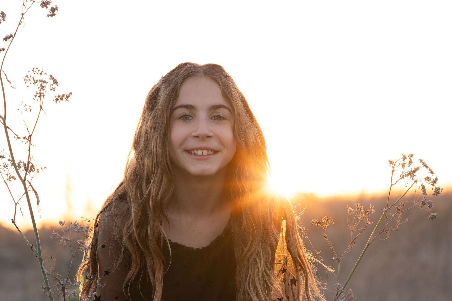 A person smiles in golden sunlight with sun flare while standing outdoors during sunset.