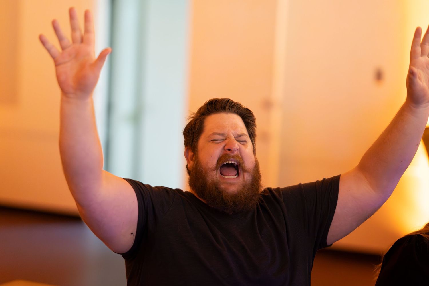 Person in black shirt raising arms enthusiastically against orange-lit background.