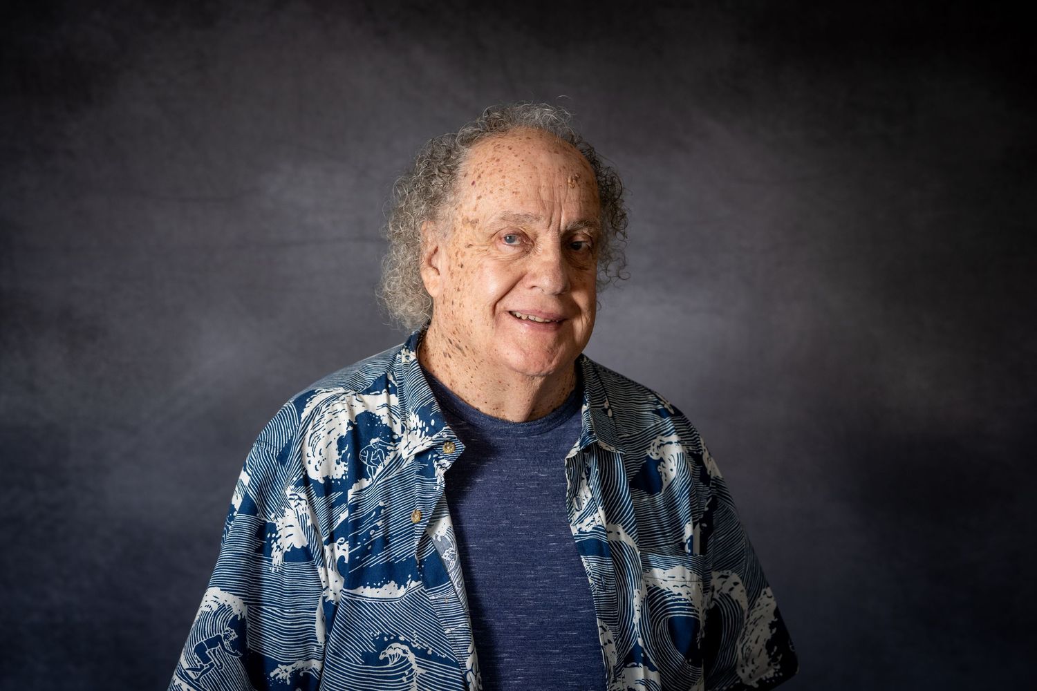 A person wearing a blue patterned shirt sits against a dark background in a candid portrait series.