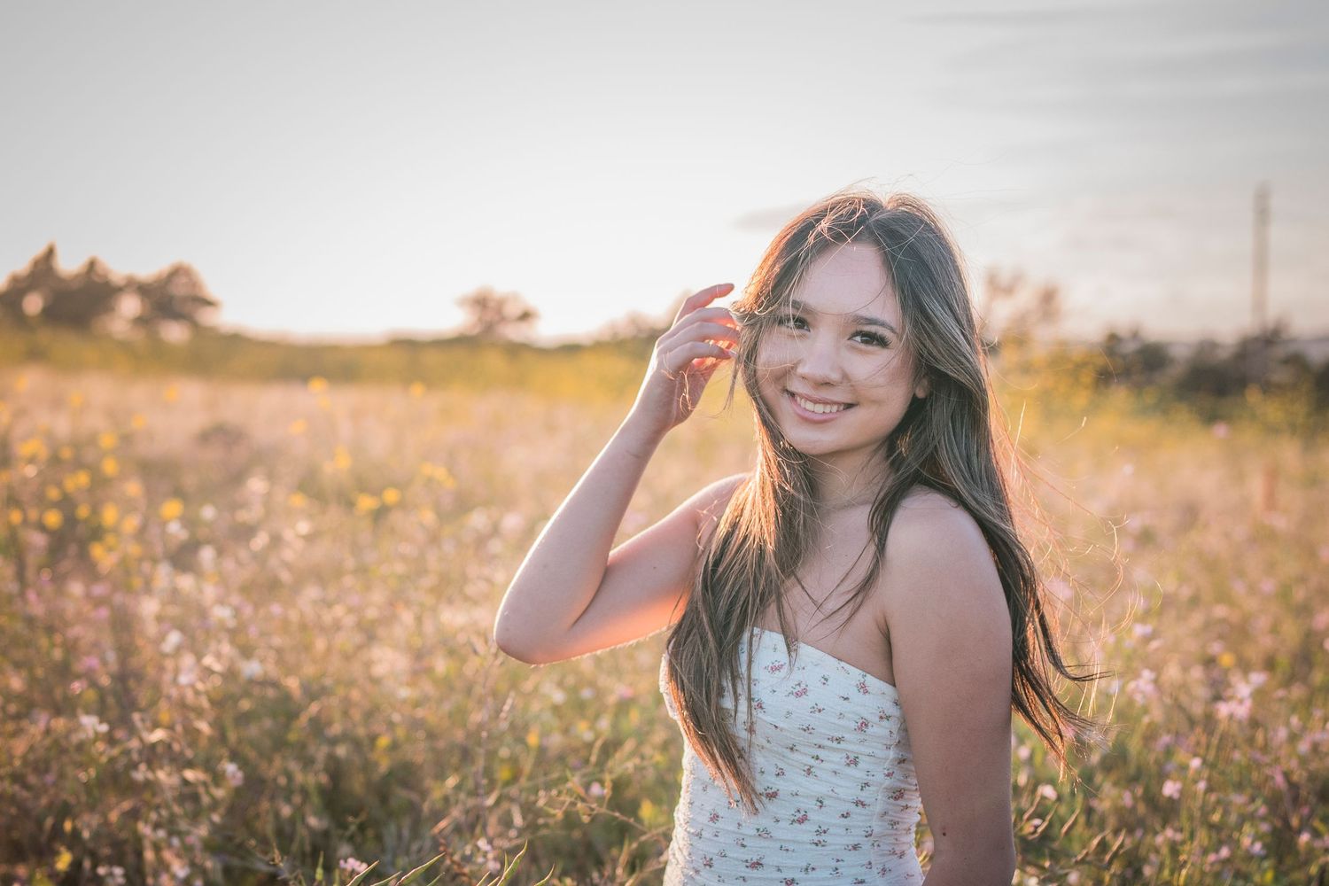Young woman smiling in a sunlit wildflower field, wearing a white dress with long brown hair blowing gently.