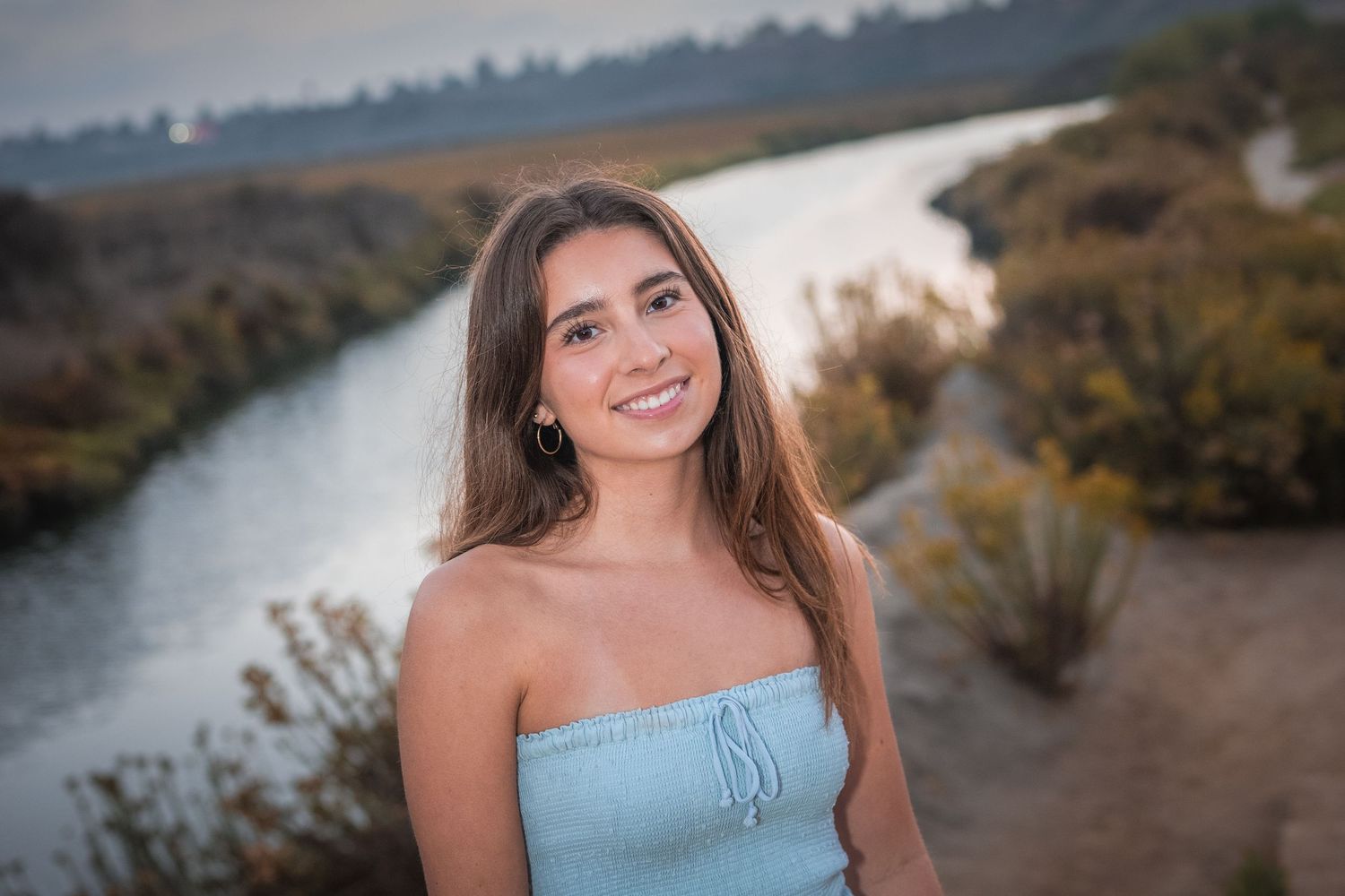 A person in a light blue strapless dress poses outdoors near a winding river at sunset.