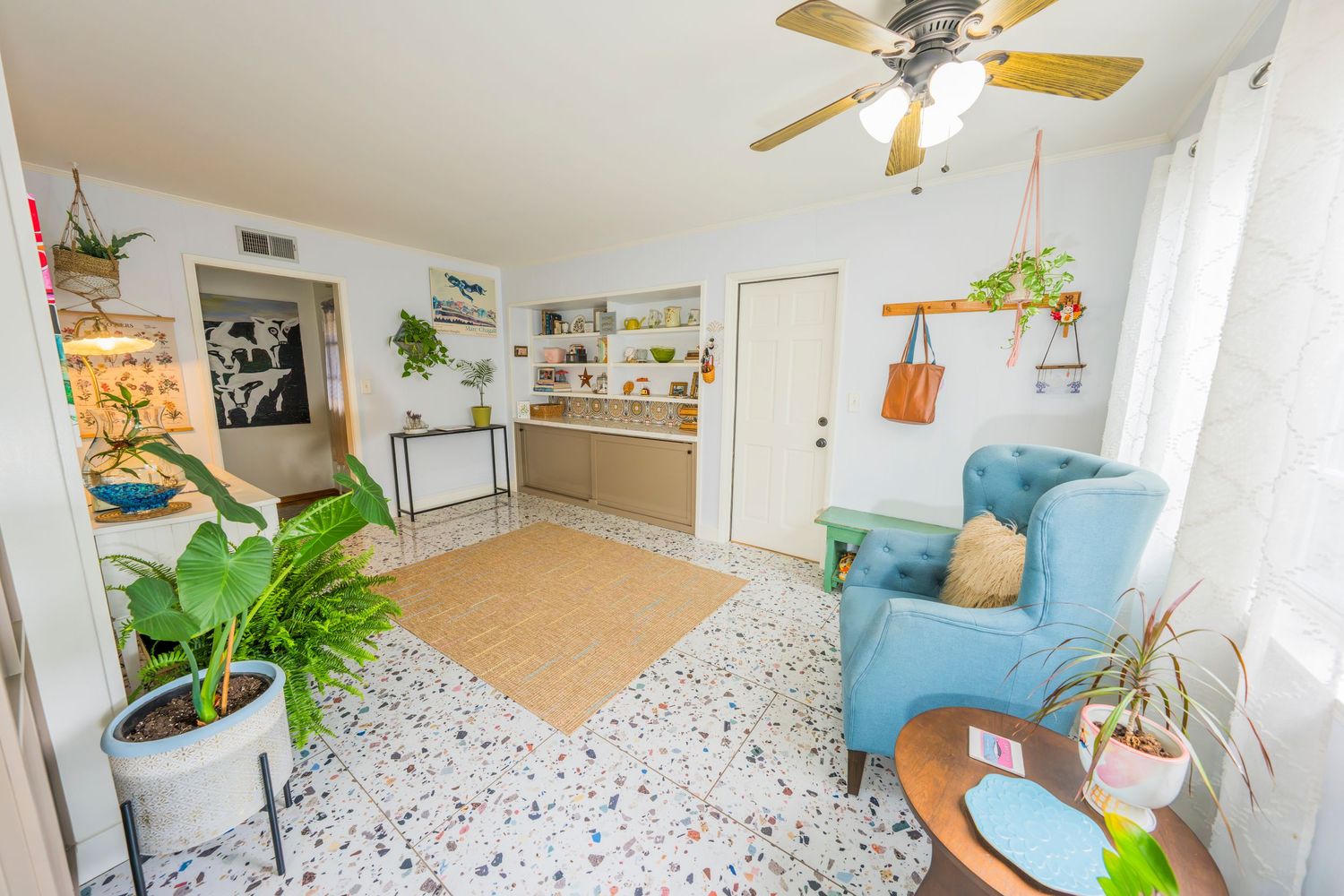 Bright living room with terrazzo floors, blue armchair, ceiling fan, indoor plants, and open shelving in background.