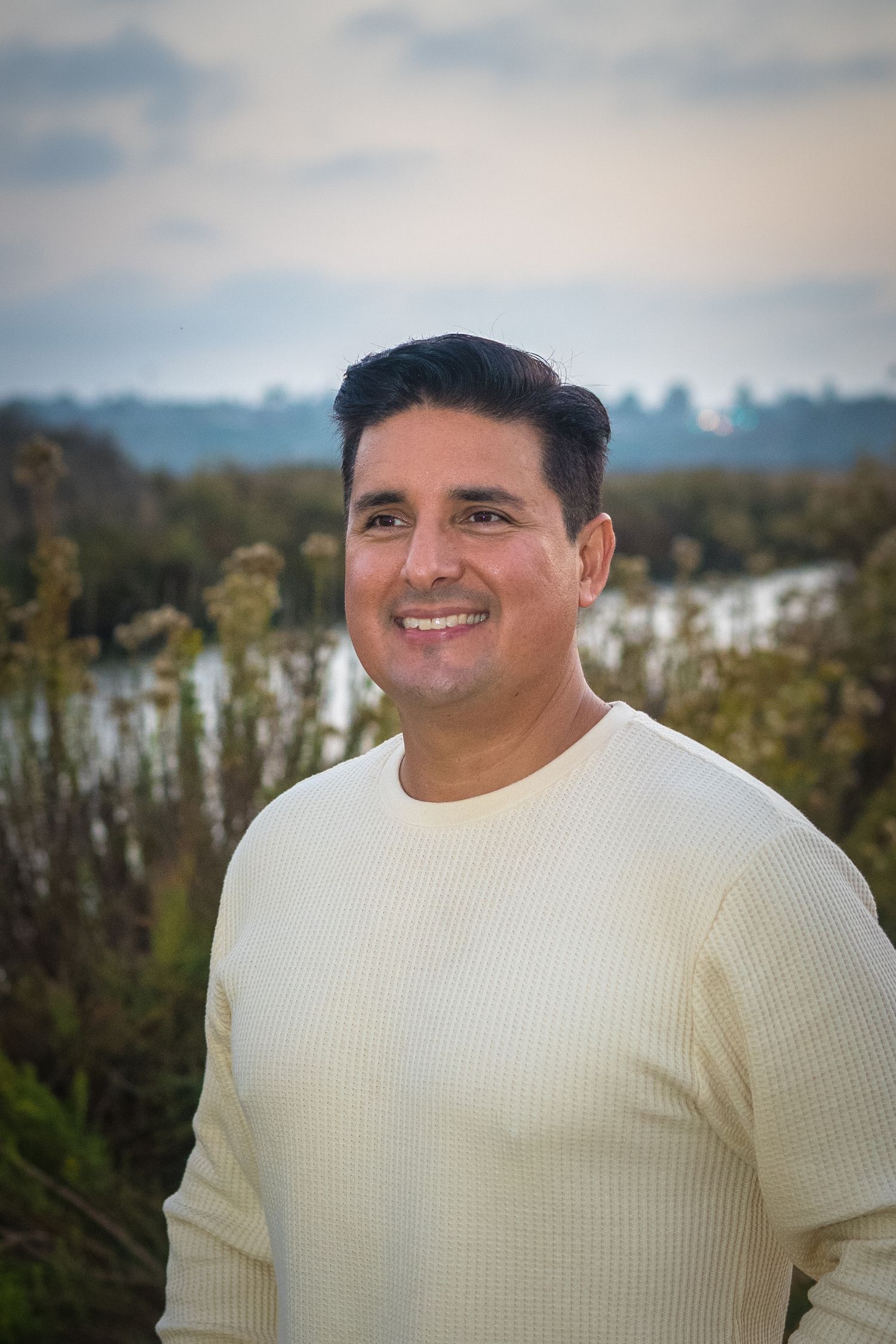 A person in a cream colored sweater smiles warmly while standing outdoors against a natural landscape background.